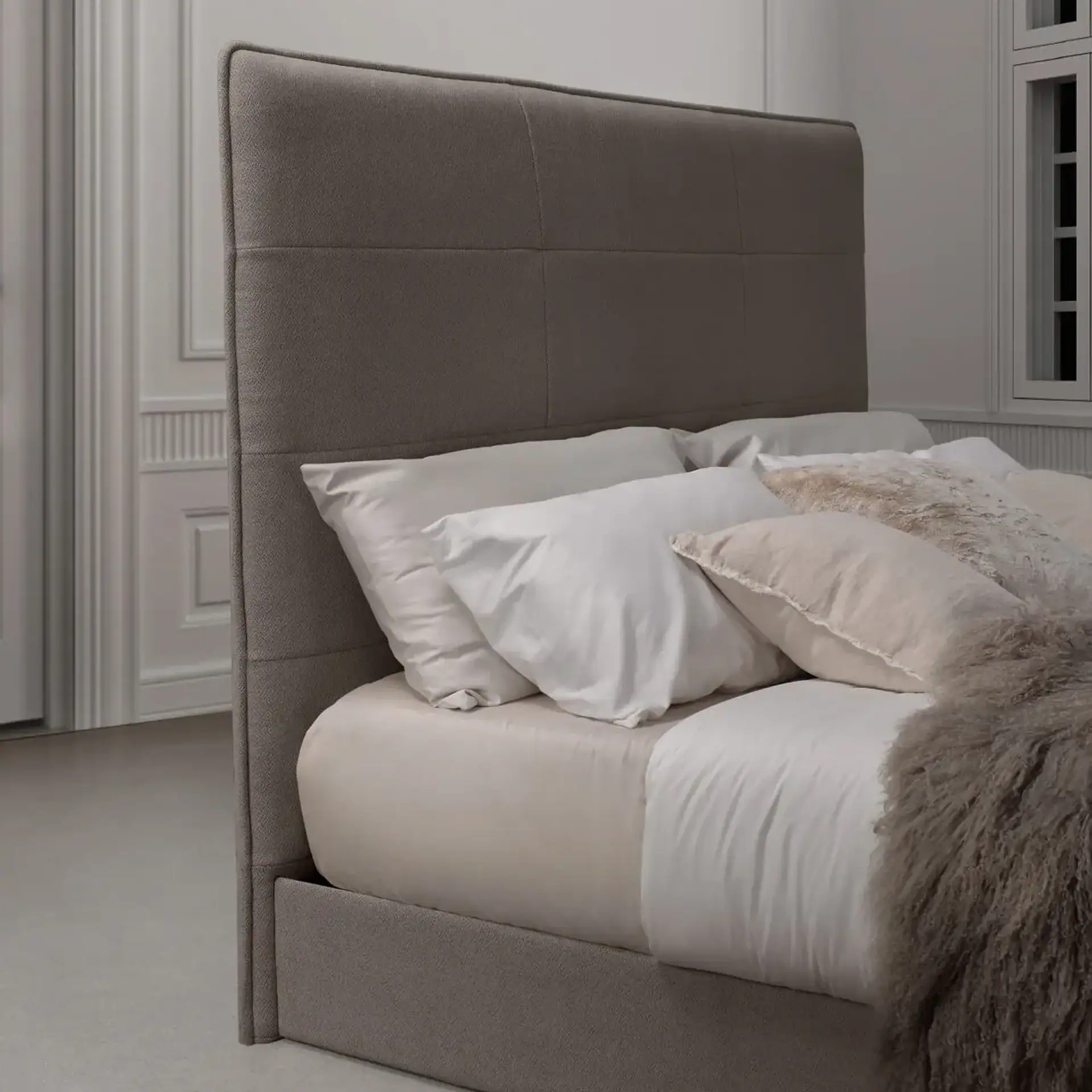 A modern bed with a tall, grey upholstered headboard and frame, dressed with white and beige pillows and a shaggy throw blanket. White architectural details are visible in the background.
