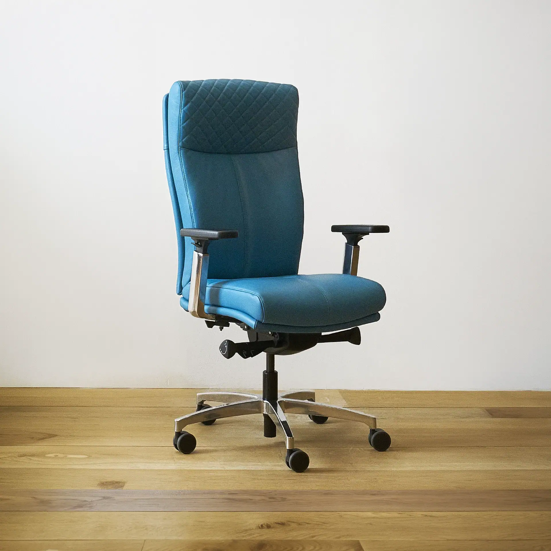 A teal office chair with a high, quilted backrest, black armrests, and a chrome five-star base with wheels, sitting on a wooden floor against a white wall.