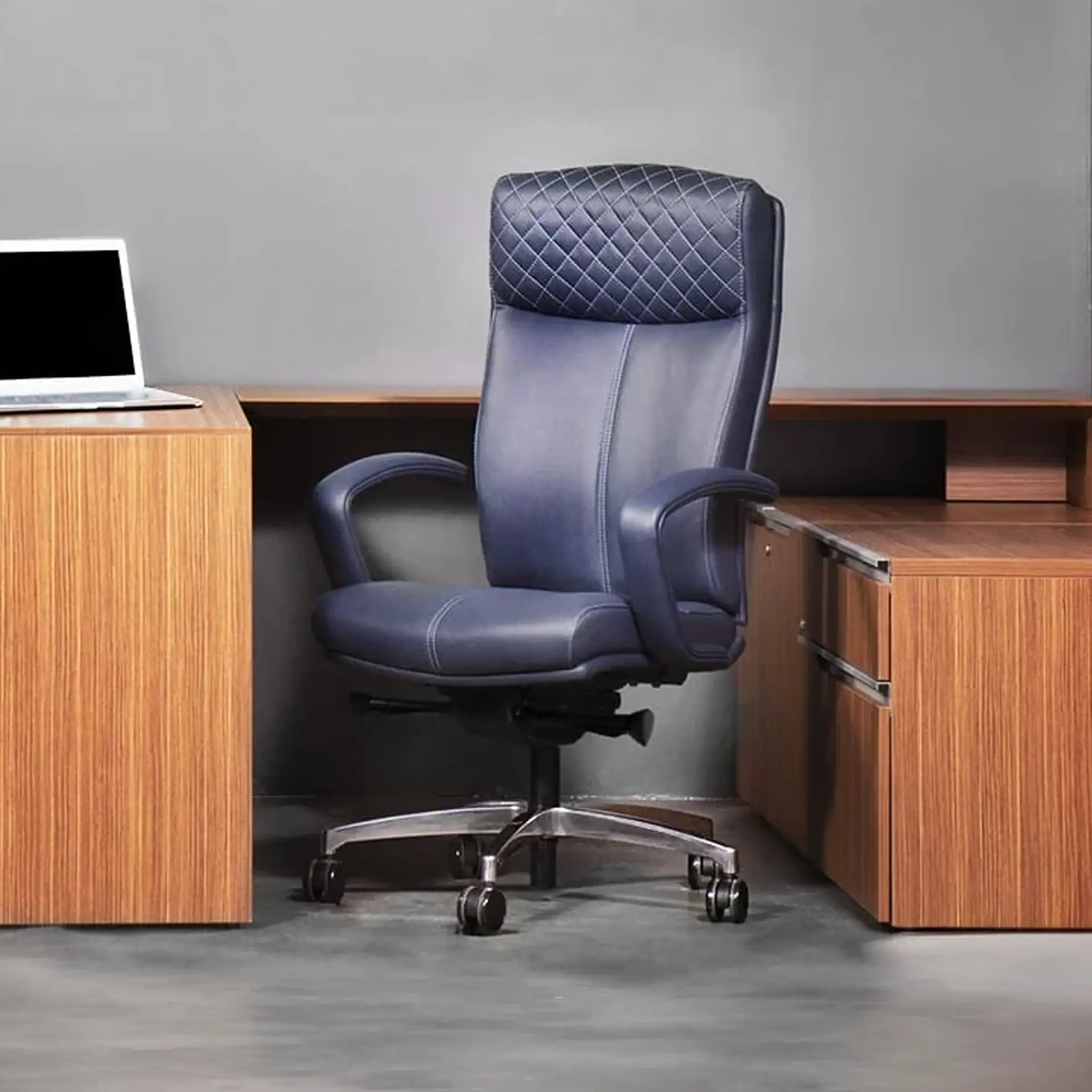A dark blue high-back office chair with a quilted headrest sits in front of a wooden desk with a laptop on it, against a gray wall.