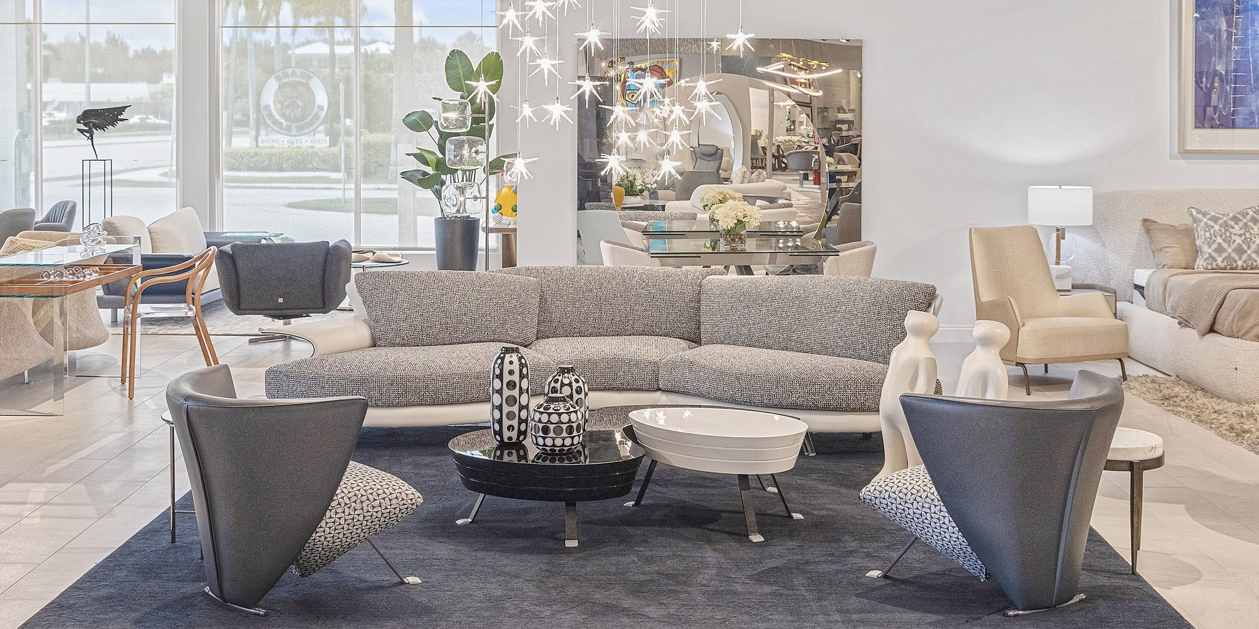 A modern furniture showroom with a grey sectional sofa, two dark grey armchairs, and two unique coffee tables on a dark rug. Brightly lit by natural light from large windows and a star-shaped chandelier, the space also features a bed, dining area, and various decor items.