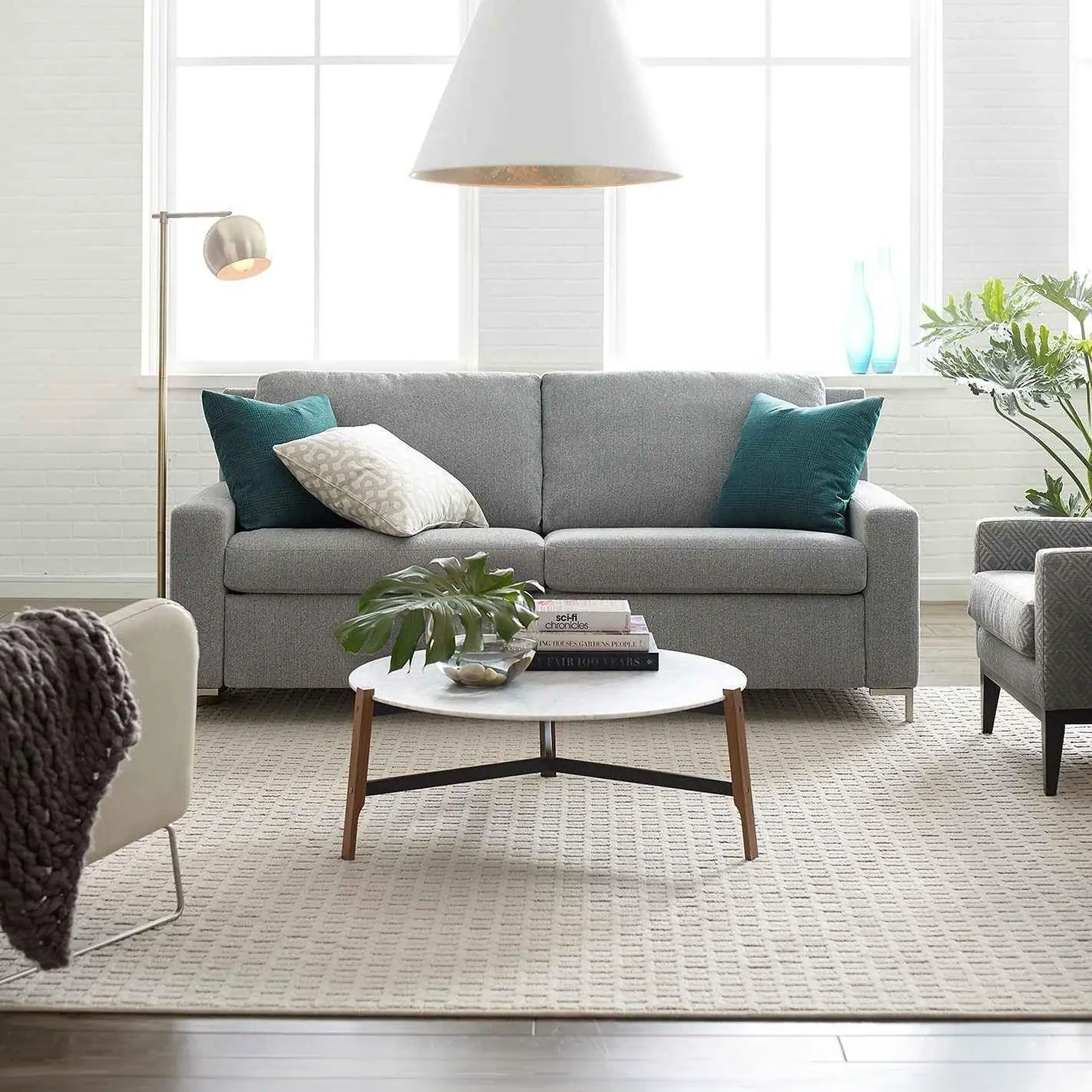 A modern living room with a gray sofa, marble coffee table, and a large window. Teal pillows and green plants add color.