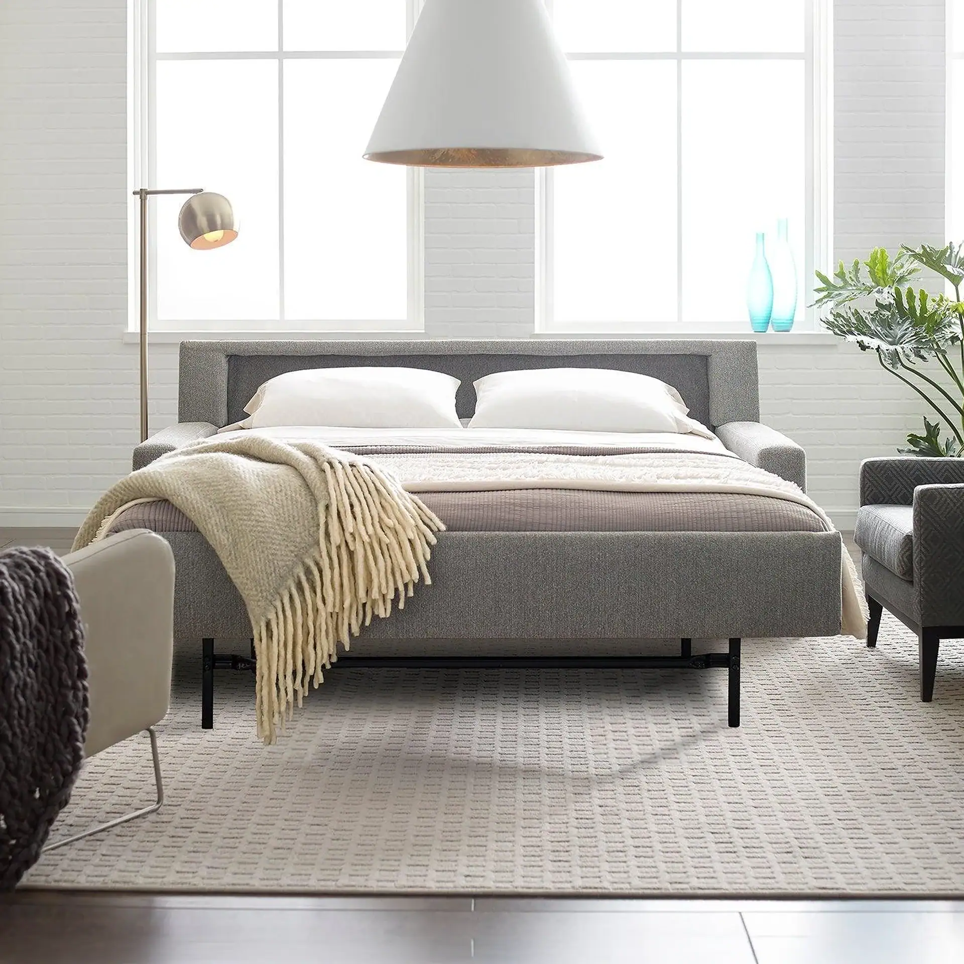 A modern bedroom with a gray upholstered bed, white pillows, and a cream throw blanket. Large windows, a white pendant light, and a brass floor lamp illuminate the space. Two armchairs, blue vases, and a plant complete the decor on a patterned rug.
