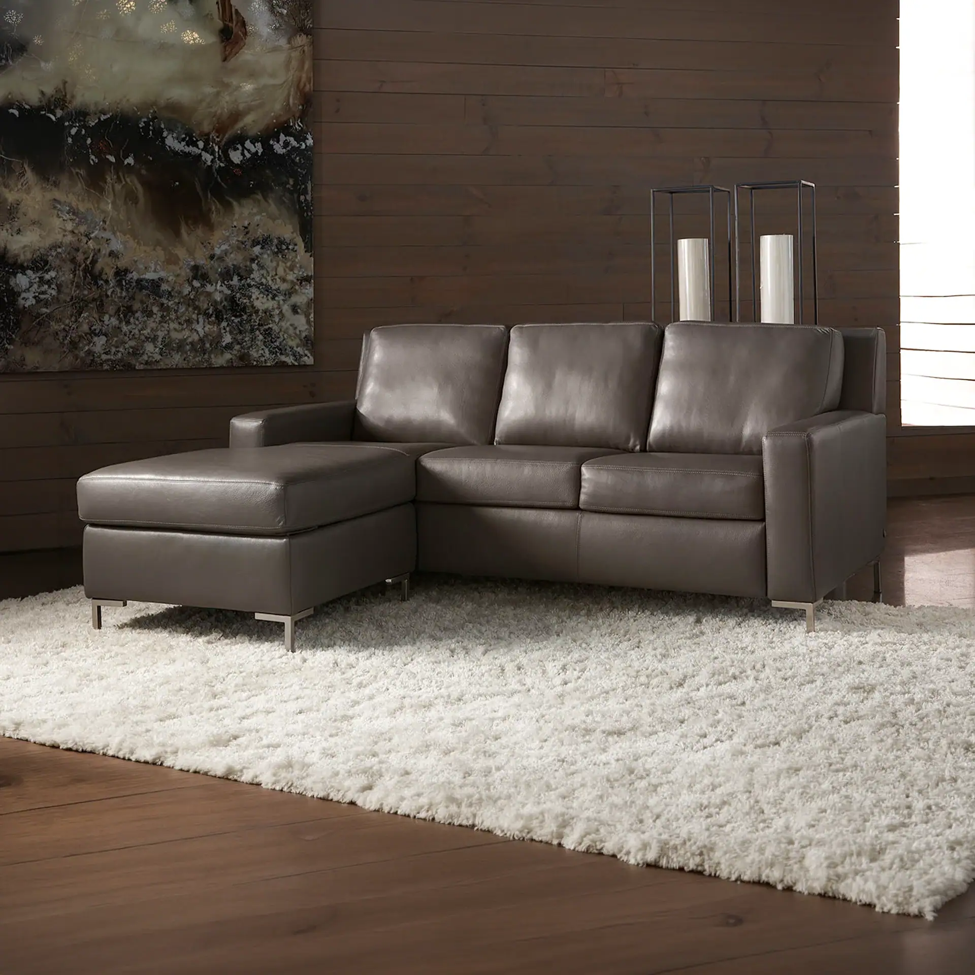A modern dark gray leather L-shaped sectional sofa with a chaise lounge sits on a white shag rug over dark wood floors. An abstract painting and two lanterns decorate the dark wood paneled wall behind it.