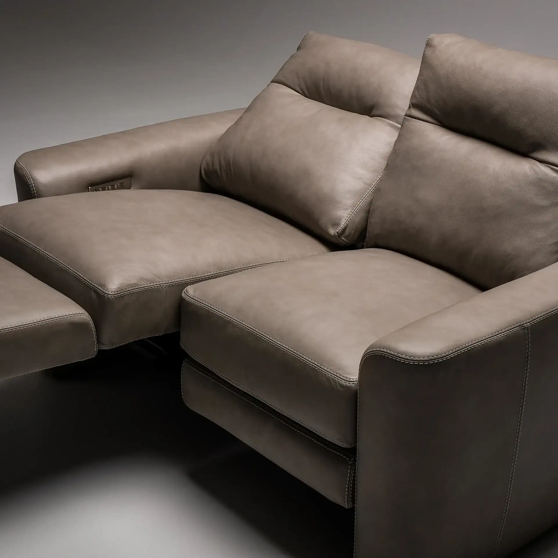 A taupe leather two-seater reclining sofa with plush cushions and white contrast stitching. The left seat is reclined with the footrest extended, showing a metallic control panel on the armrest.