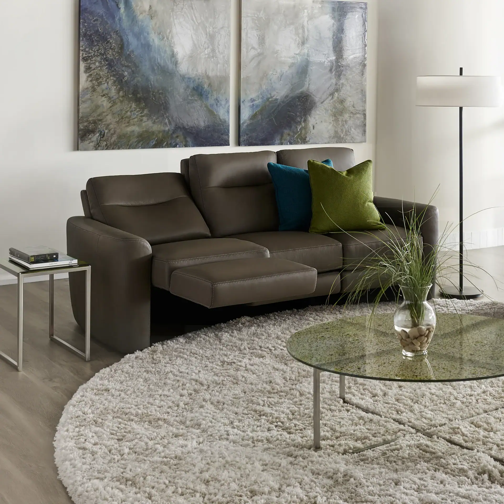 A brown leather reclining sofa with teal and green pillows, a cream shag rug, glass coffee table with a plant, and abstract art in a modern living room.
