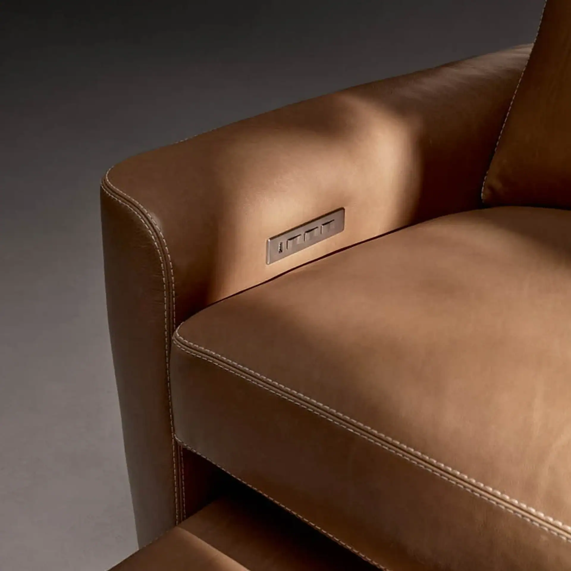 Close-up of a brown leather armchair with white stitching and an embedded metallic control panel on the armrest.