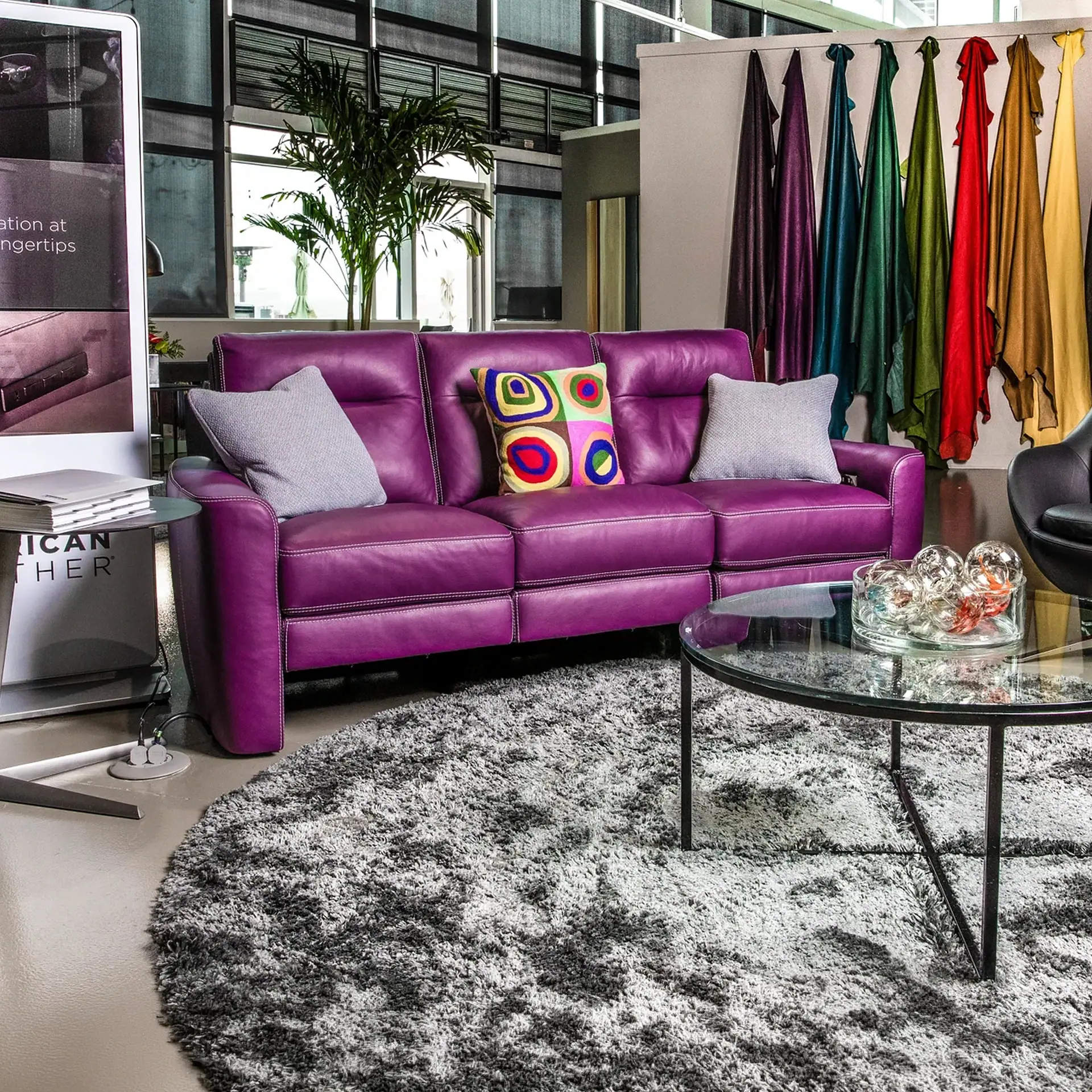 A purple leather sofa with colorful pillows, a glass coffee table, and a gray shag rug in a modern showroom.