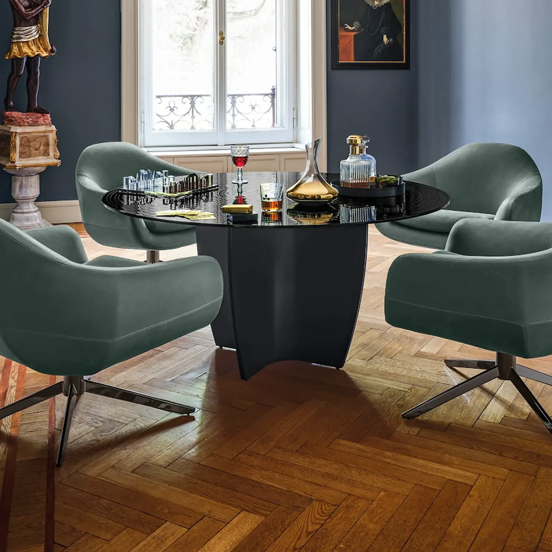 An interior room with dark walls, a herringbone wood floor, a round black table, and four dark green chairs. The table has a modern chess set, decanters, and drinks. A statue and a framed painting decorate the room.