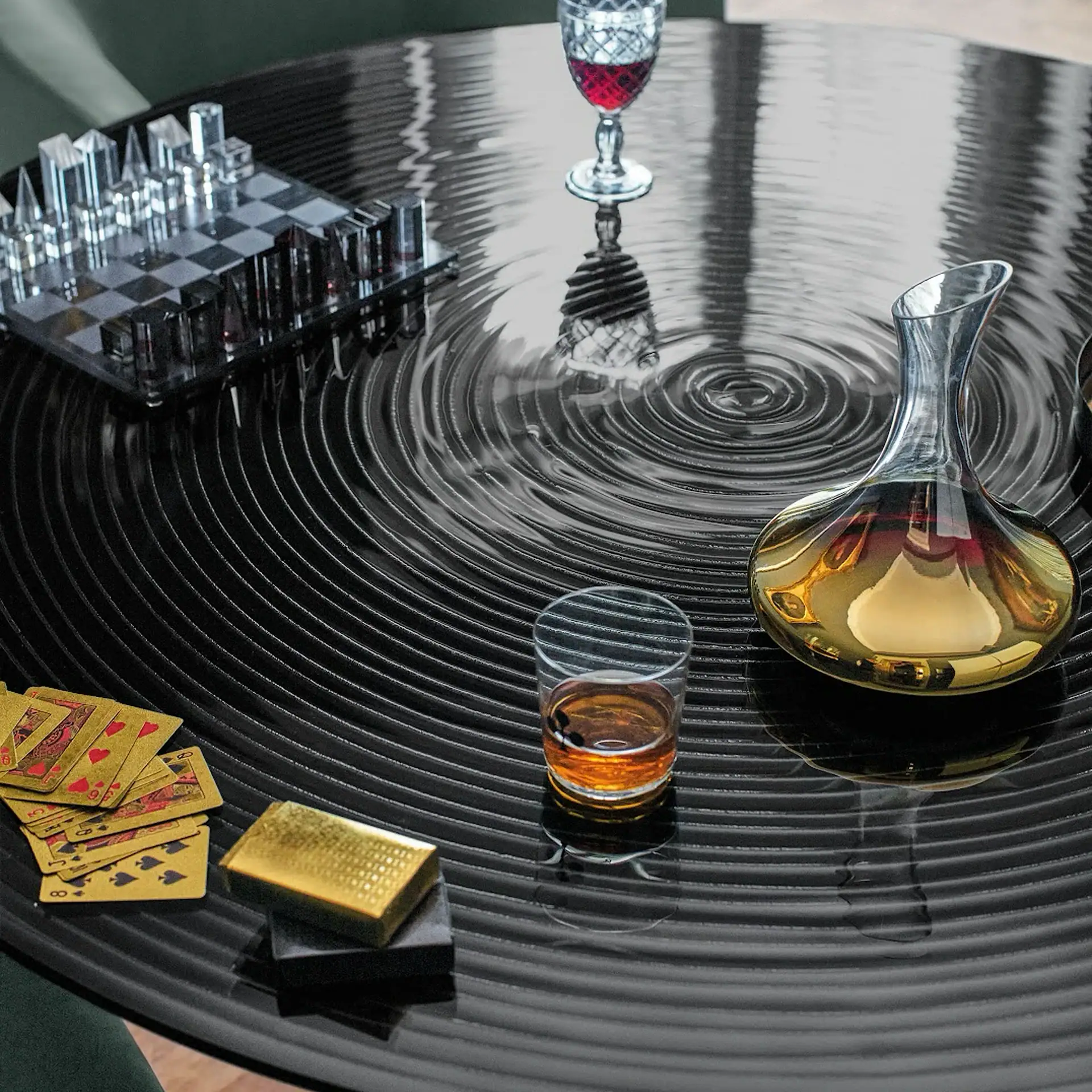 A black, glossy, rippled table holds a clear chess set, a decanter with amber liquid, a wine glass with red liquid, a shot glass, and gold playing cards with a matching card box.
