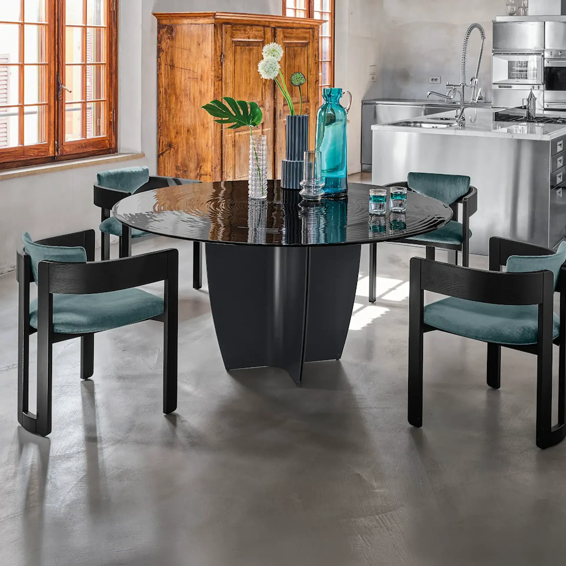 A modern dining room with a black round table, teal velvet chairs, and a stainless steel kitchen island in the background.