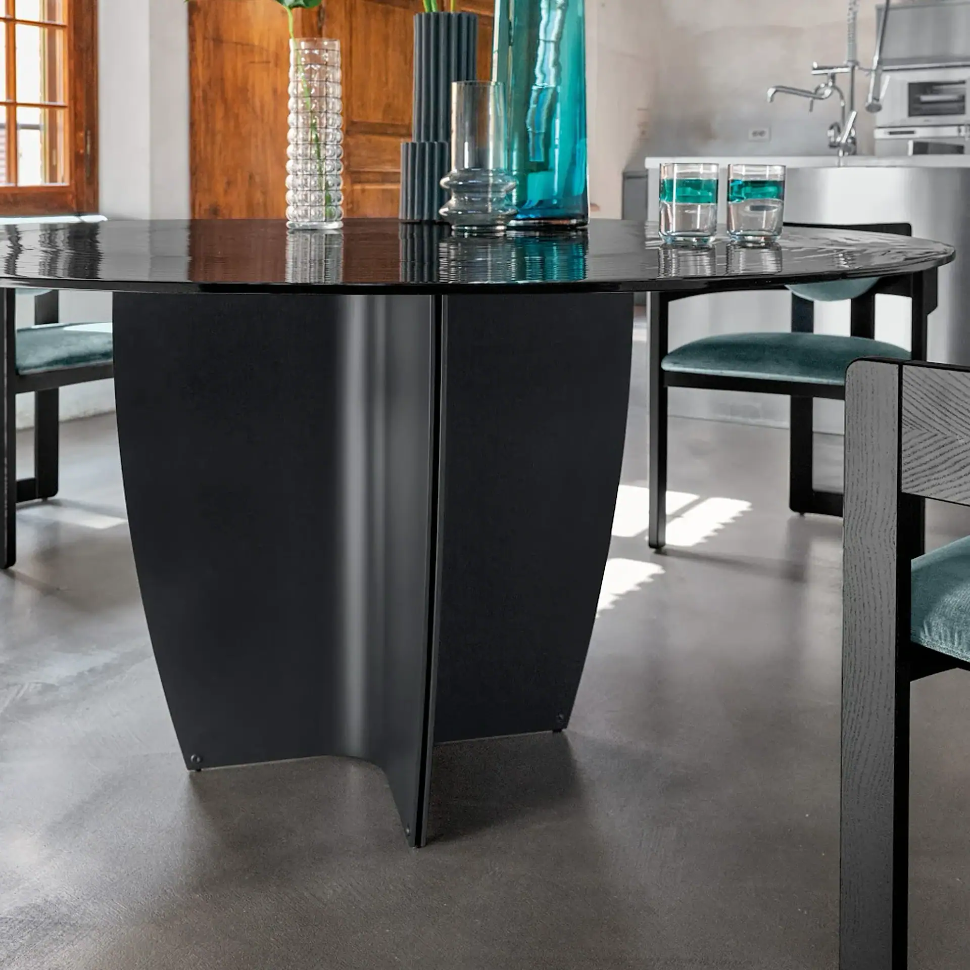 A modern dining room with a round, glossy black table and sculptural base, surrounded by black chairs with teal velvet seats. Vases and glasses adorn the table, with a kitchen and wooden wall in the background.