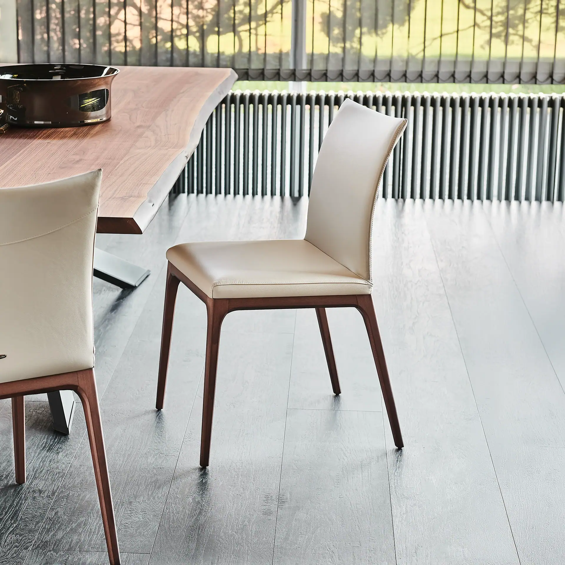 A modern dining room with a live-edge wooden table, two cream leather chairs, and a large window with vertical blinds.