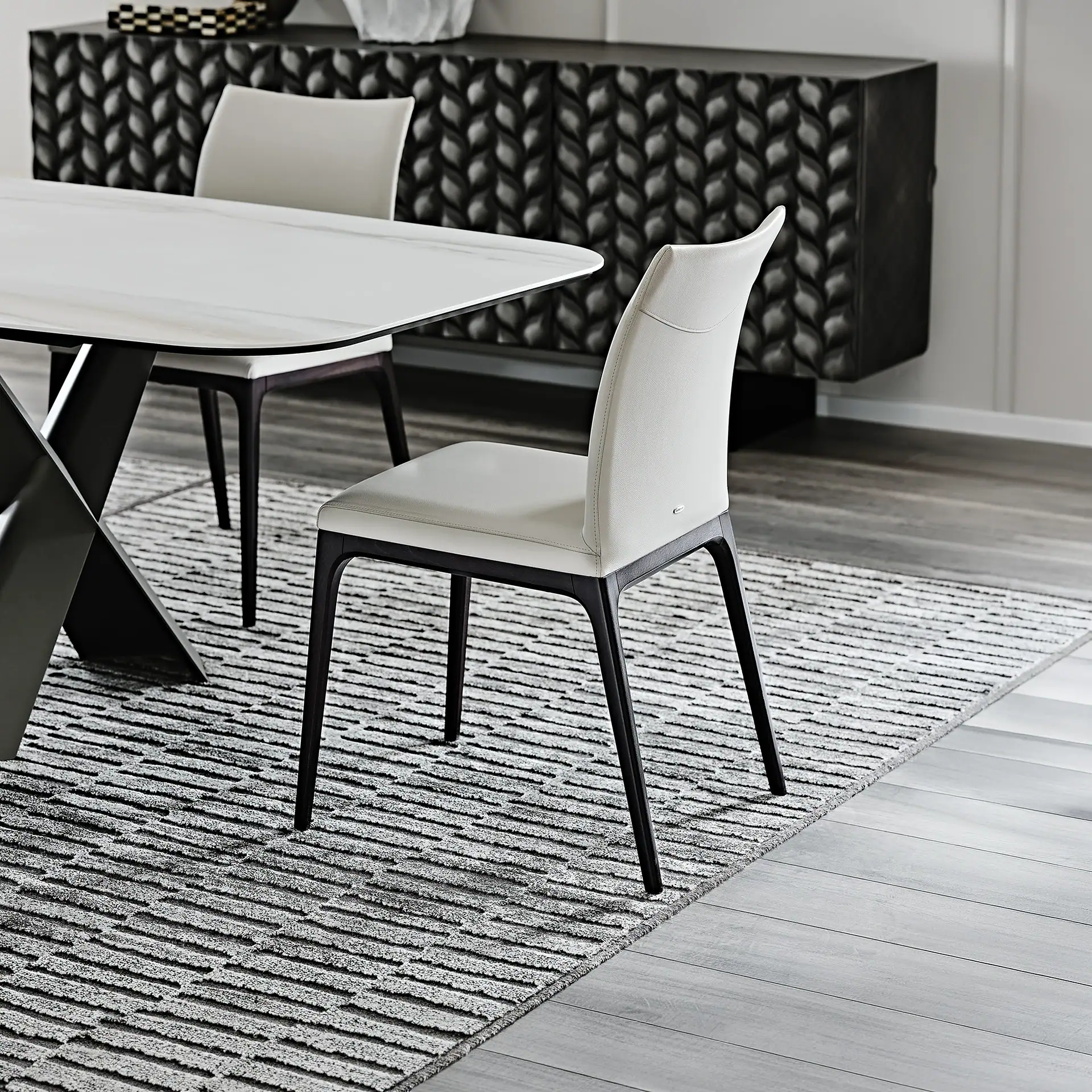 A modern dining room with a white marble-look table, light upholstered chairs, and a dark textured credenza on a grey striped rug.