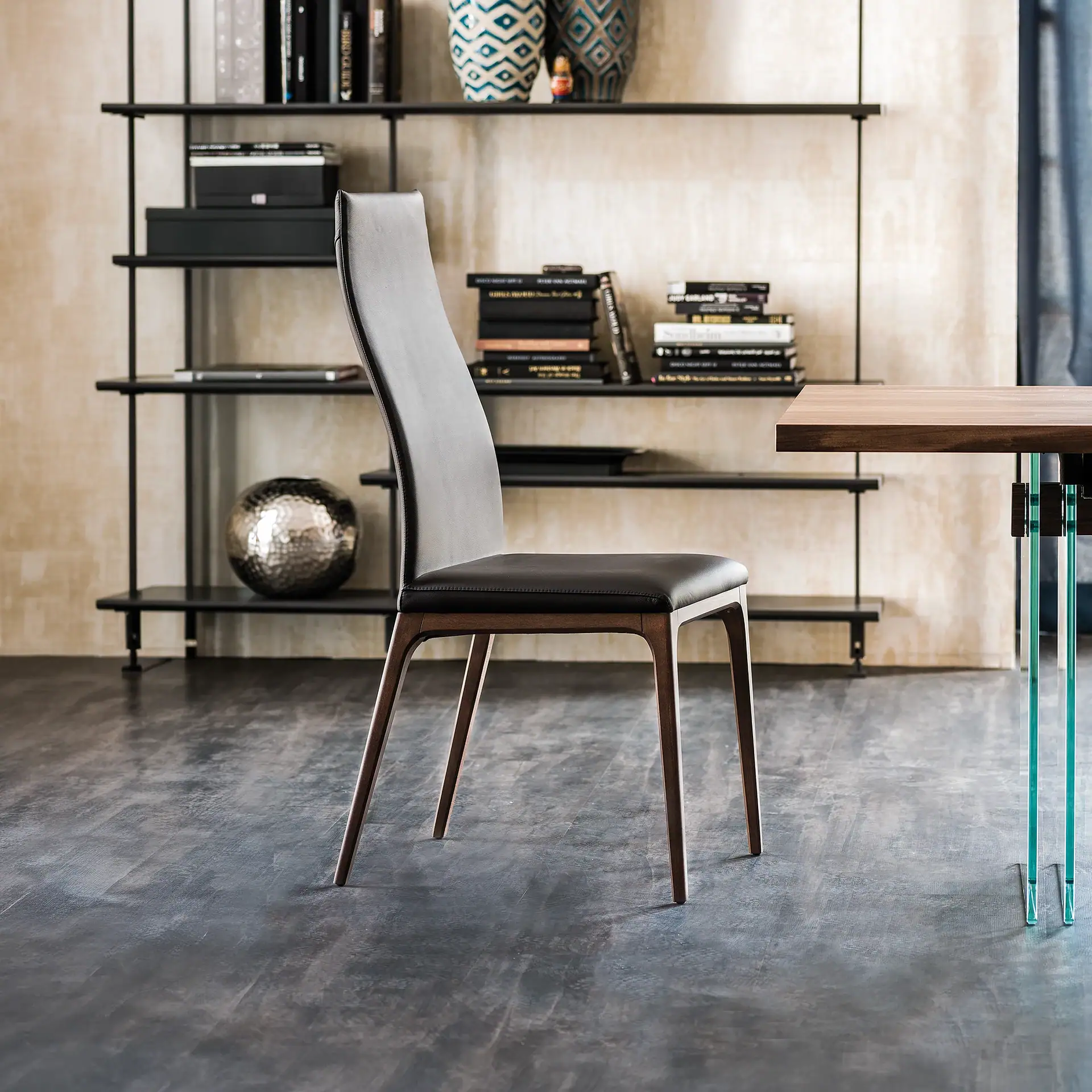 A modern dark gray leather dining chair with wooden legs, in a room with a black metal shelving unit filled with books and decor, and a wooden table with glass legs.