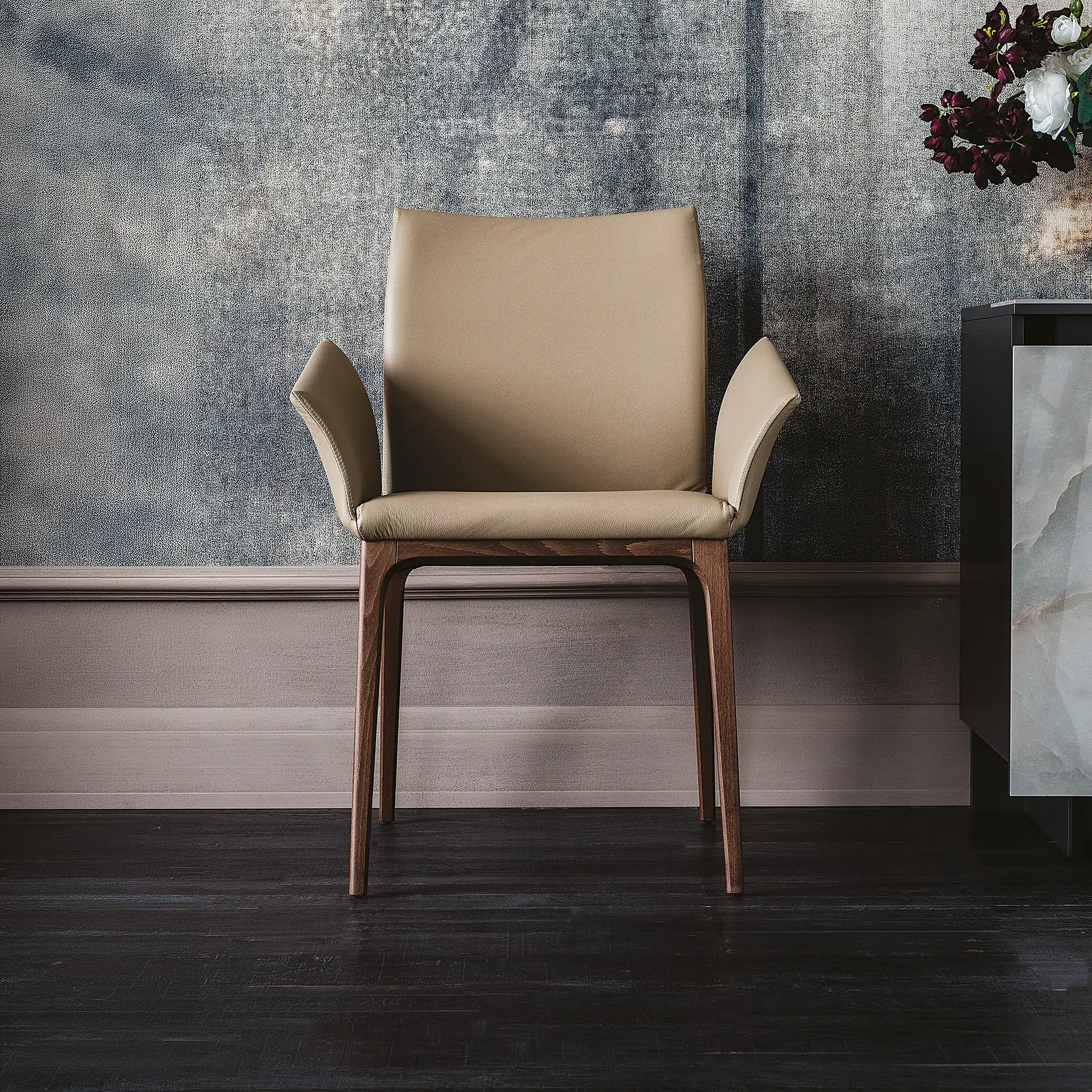 A modern beige leather chair with a dark wooden frame sits against a blue-gray textured wall. Dark wood floors are visible below, and a cabinet with flowers is partially seen on the right.