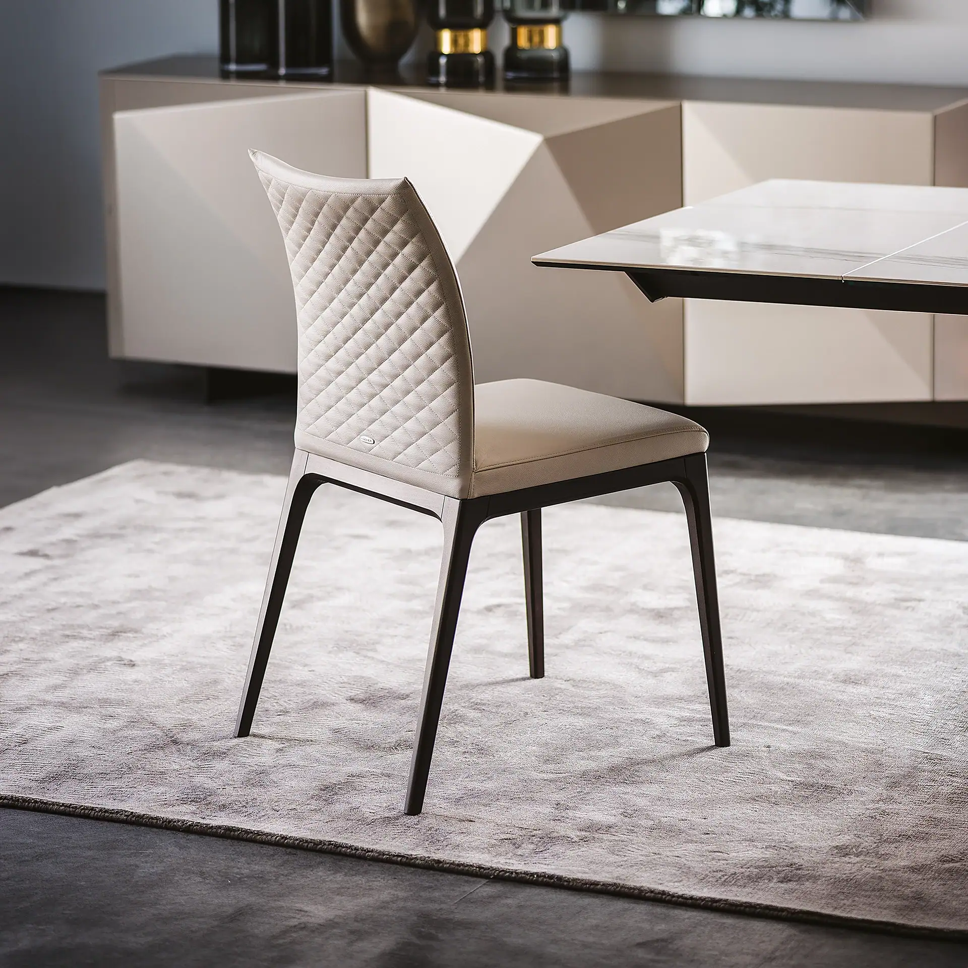 A light beige dining chair with a quilted backrest and dark legs sits on a gray rug, next to a table and a geometric sideboard.