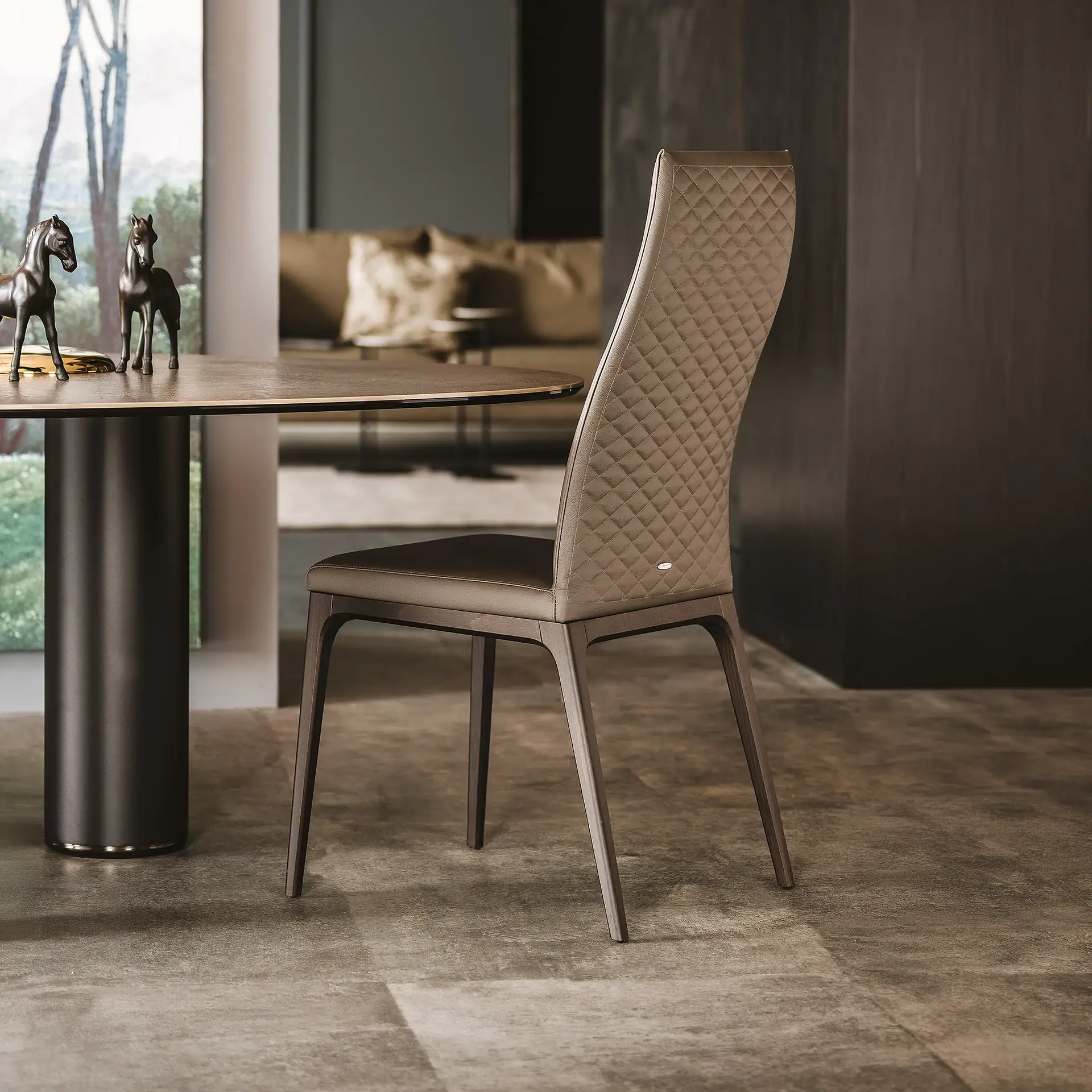 A taupe quilted dining chair next to a round dark table with two horse figurines, in a modern interior with a large window.