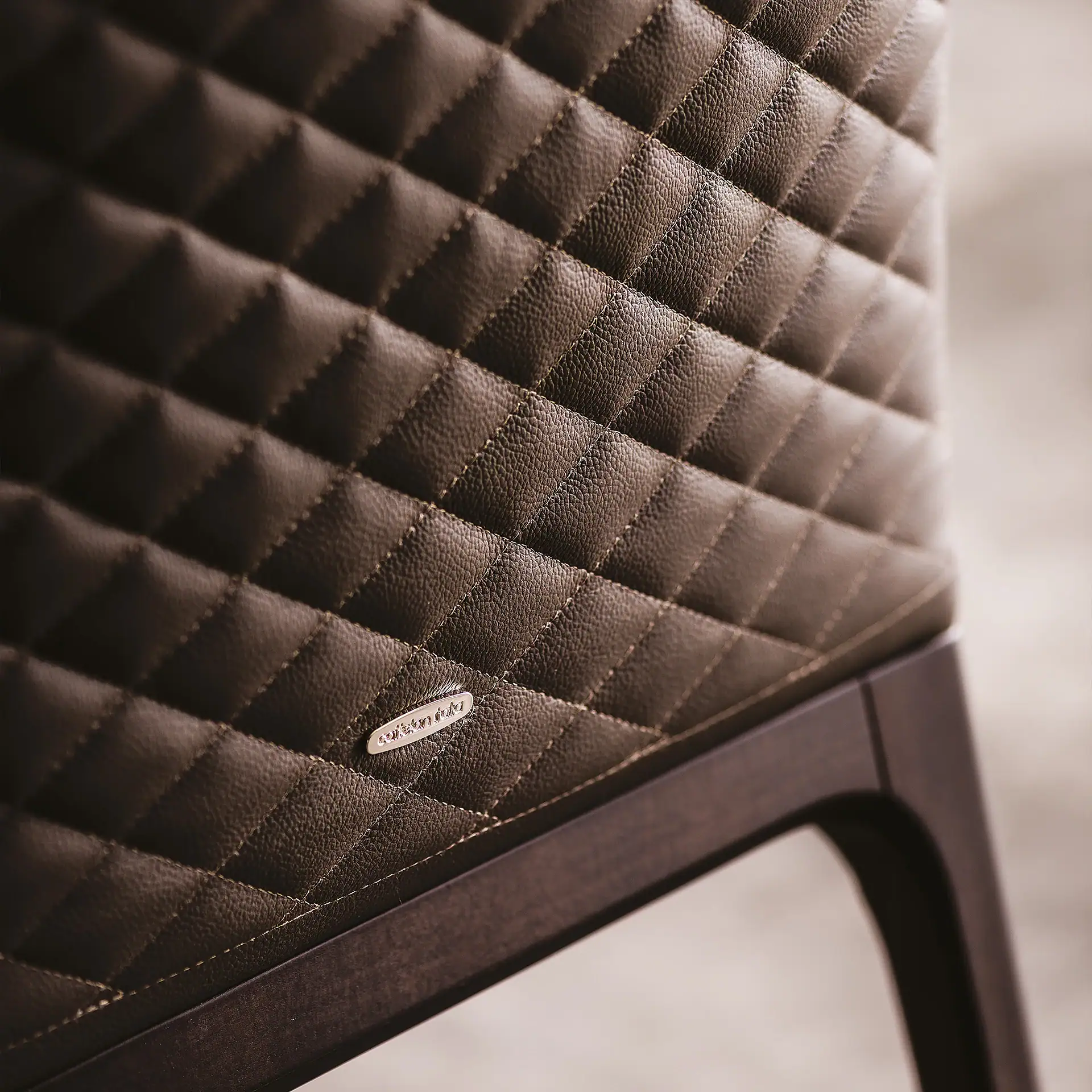 Close-up of dark brown quilted leather upholstery with diamond stitching, a metallic logo, and a wooden frame.
