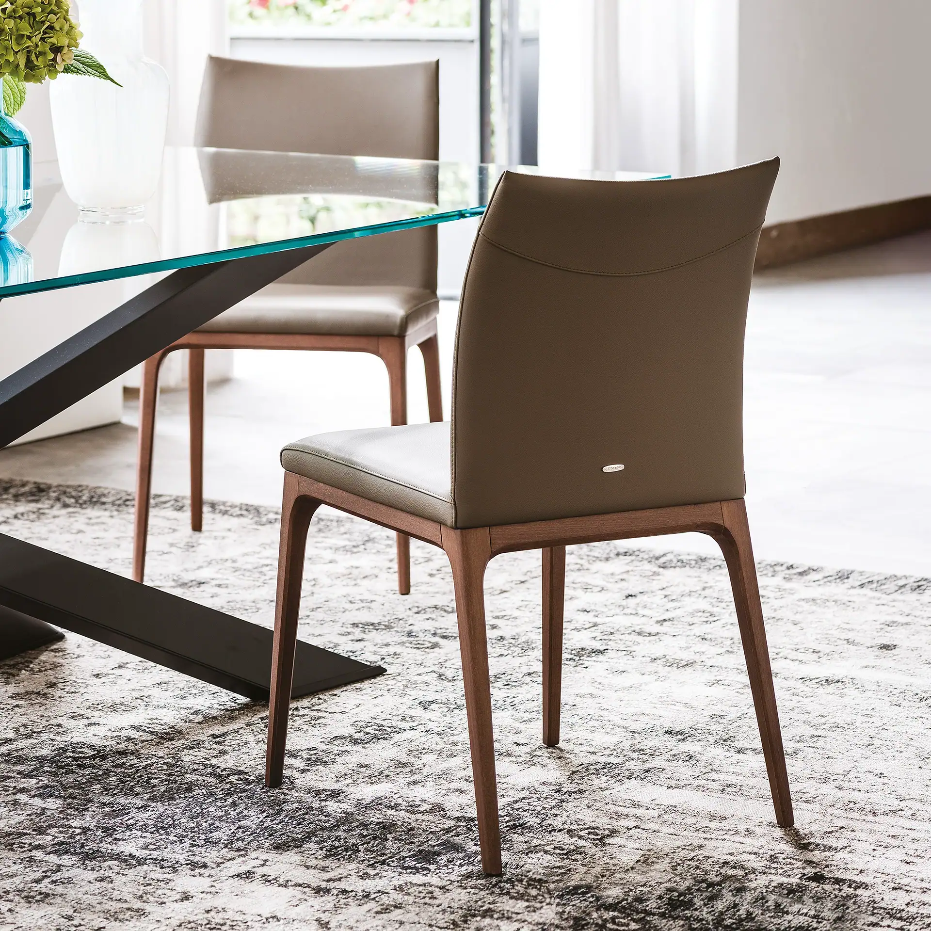 A modern dining chair with olive-green upholstery and wooden legs, seen from the back, next to a glass-top dining table on a patterned rug.