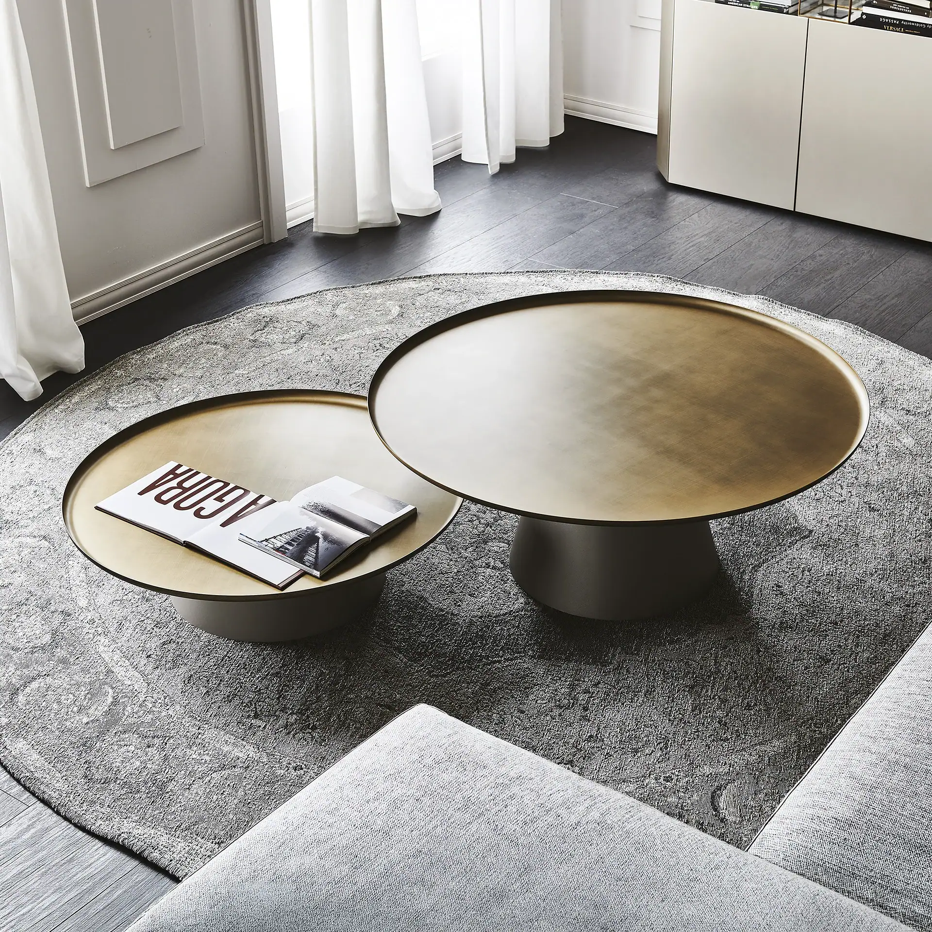Two round coffee tables with brass tops on a grey patterned rug, with a magazine on the smaller table. A grey sofa and white curtains are visible.