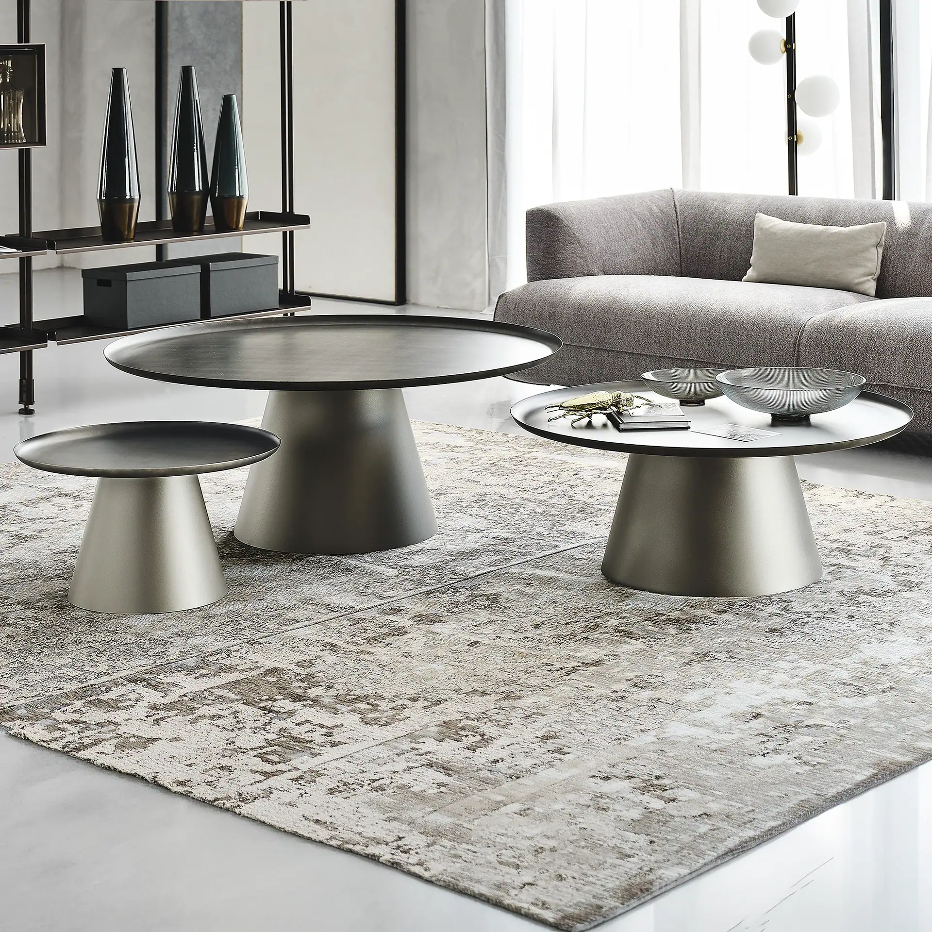 Three modern round metal coffee tables with conical bases are arranged on an abstract patterned rug in a contemporary living room. A gray sofa and shelving unit are in the background.