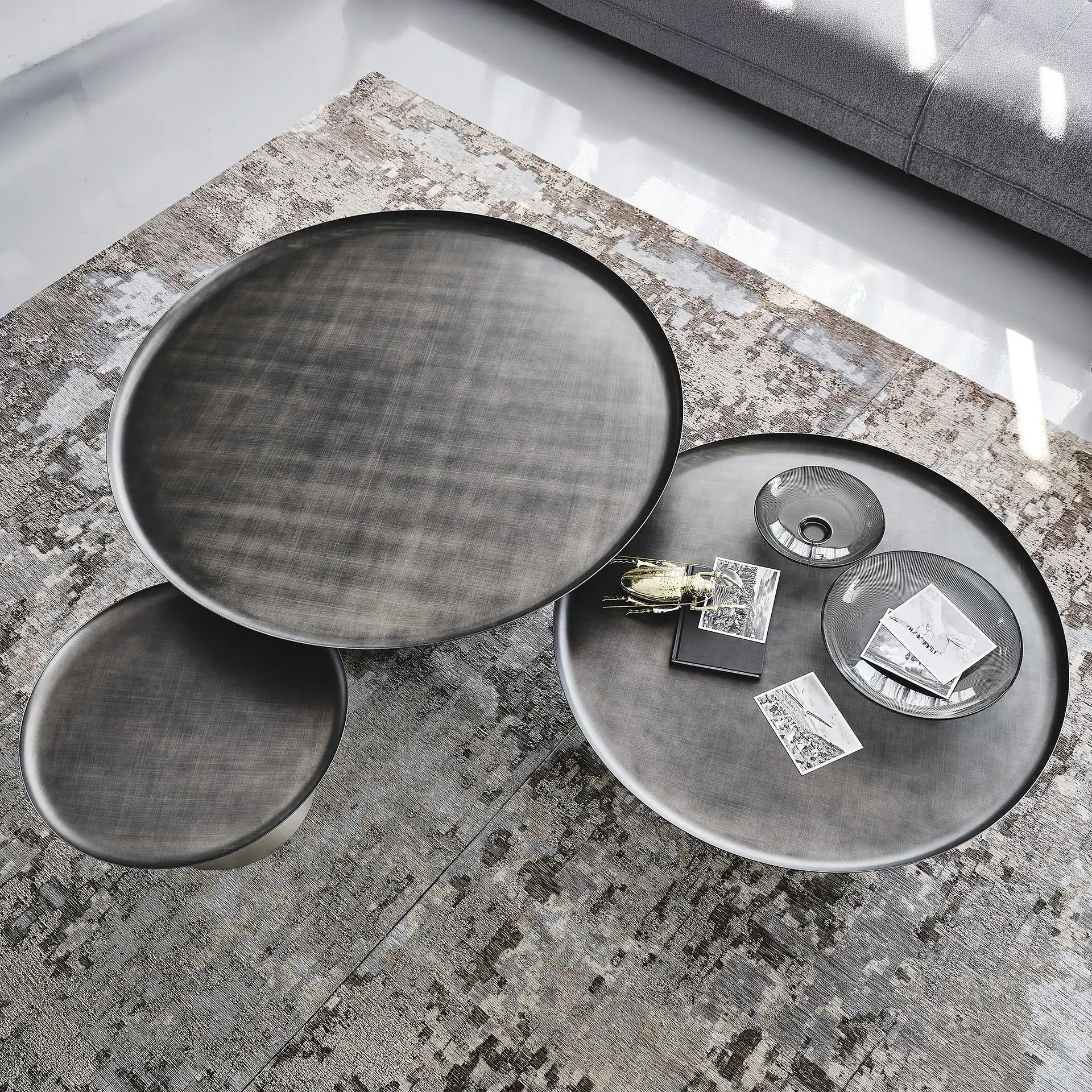 Overhead view of three round, dark grey metallic coffee tables on a patterned rug. The tables are layered, with one holding a golden beetle, a small book, black and white photos, and two ribbed glass bowls.