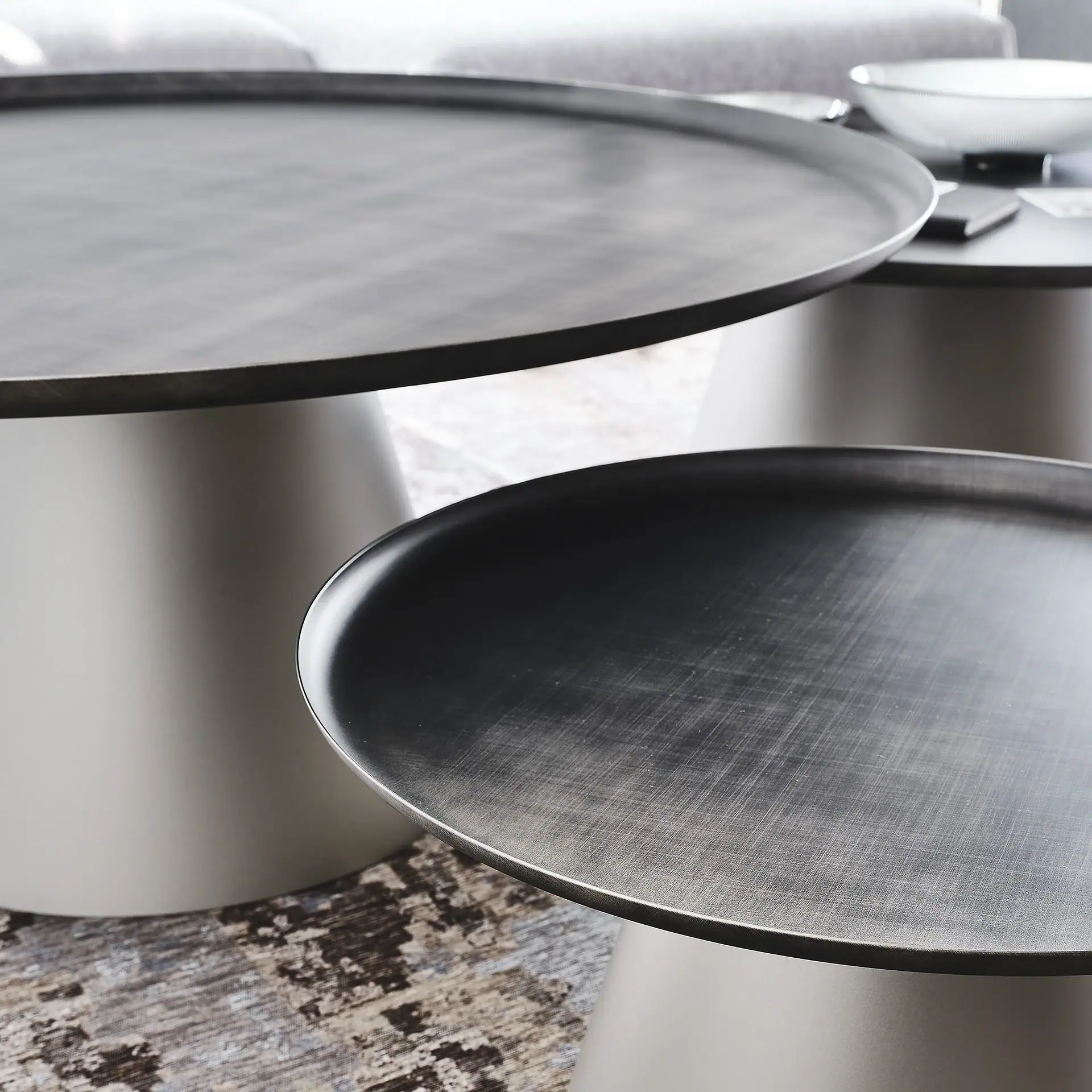 Two modern round coffee tables with dark metallic tray tops and light gray cylindrical bases, set on a patterned rug in a living room.