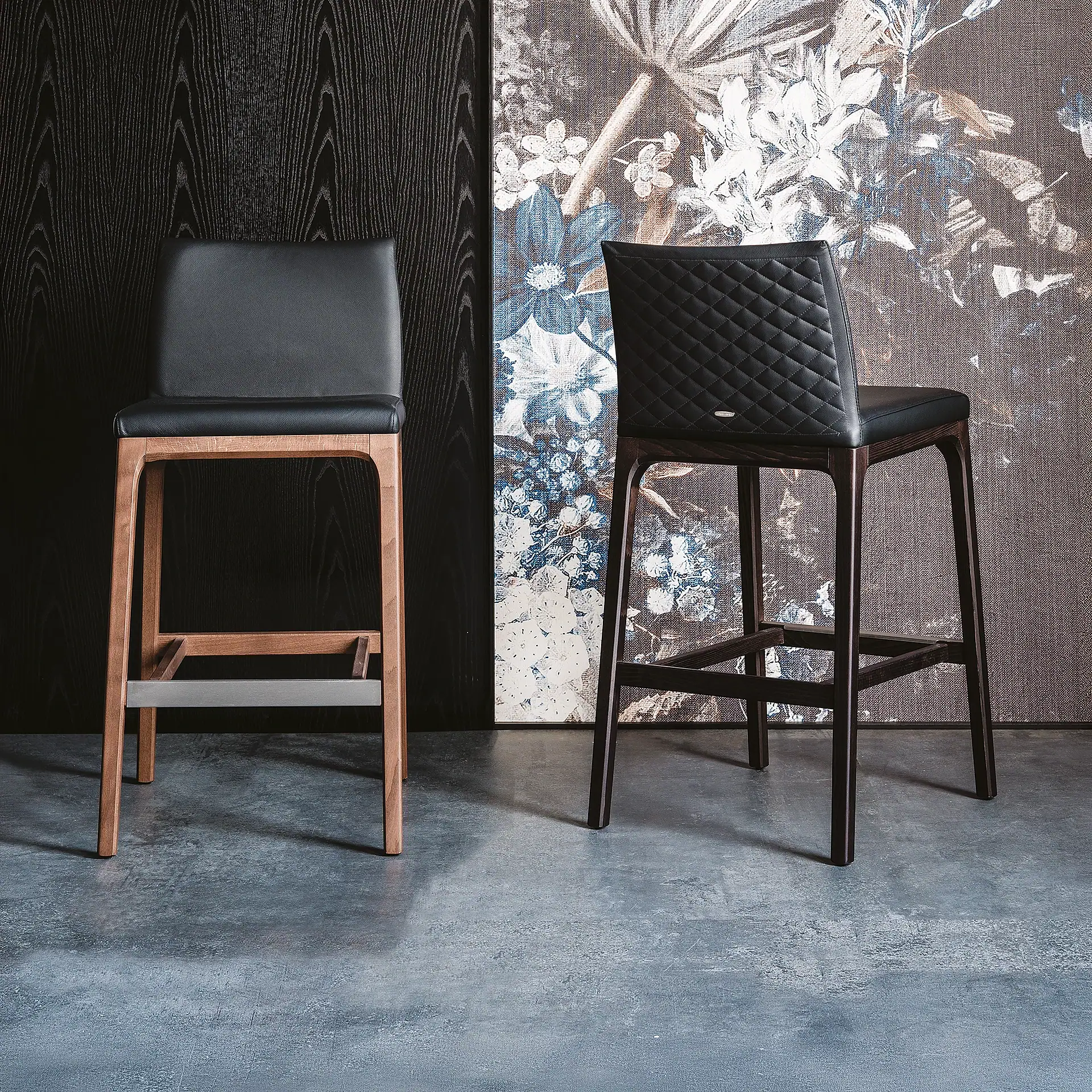 Two bar stools, one light wood with smooth black leather, one dark wood with quilted black leather, against a dark wood and floral patterned background.