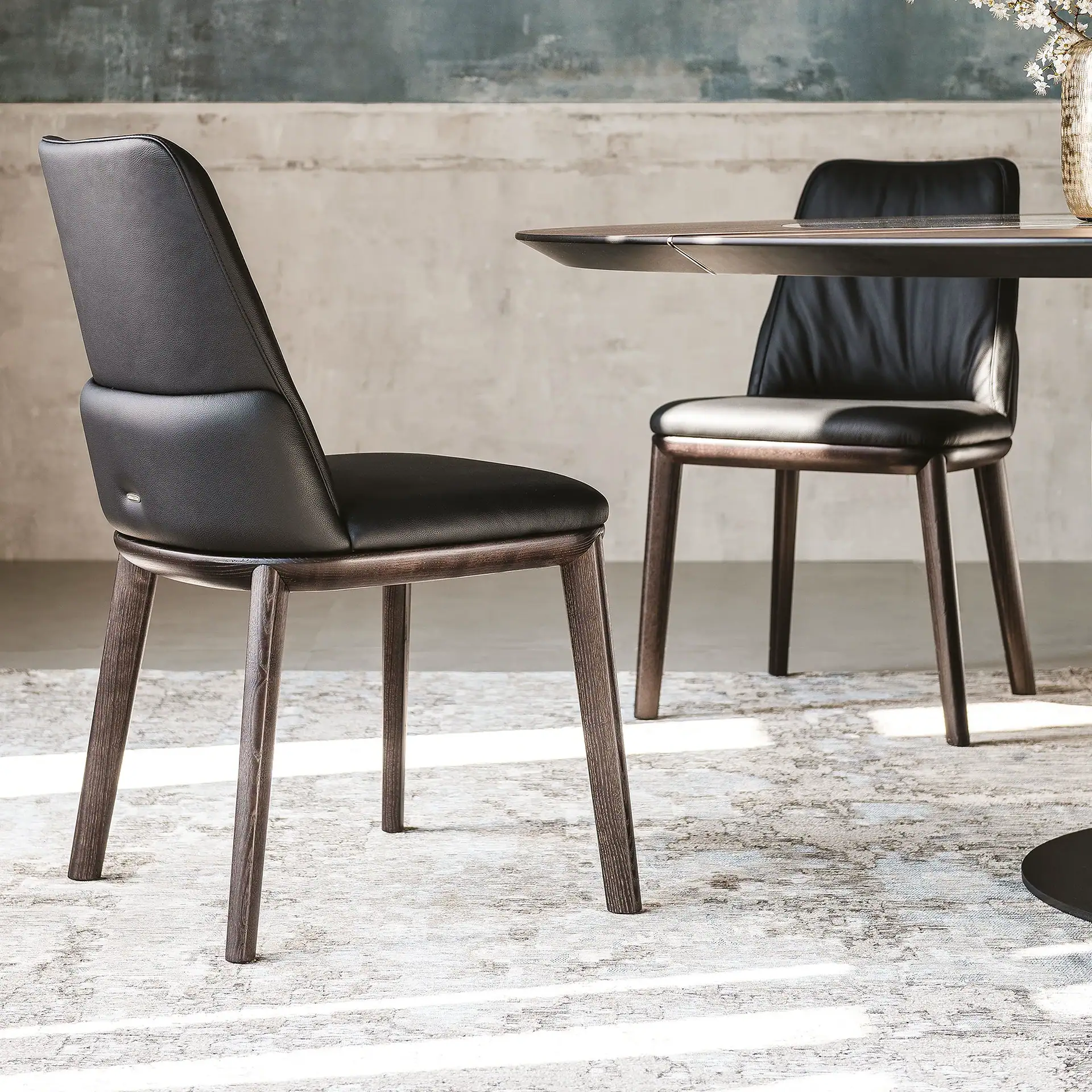 Two black leather dining chairs with dark wooden frames and a round dining table on an abstract patterned rug, against a textured grey wall.