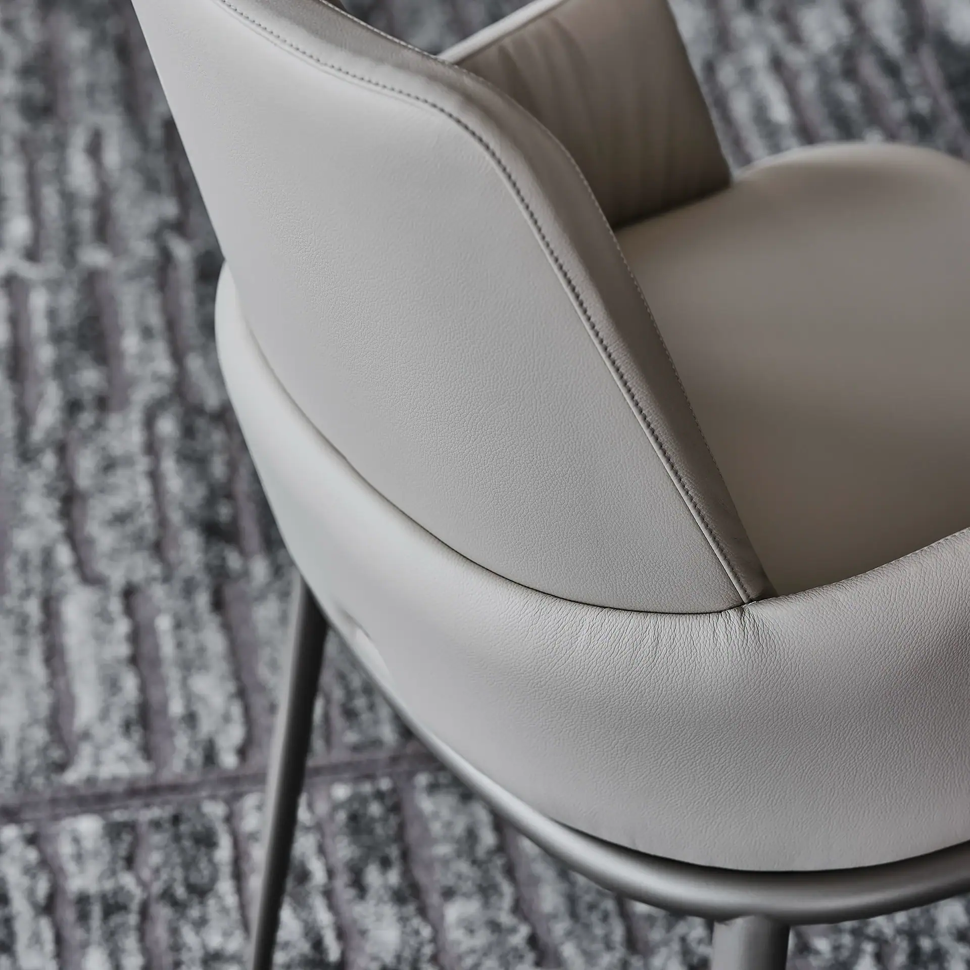 Close-up of a light gray upholstered chair with visible stitching and a metallic frame, set against a blurred dark patterned rug.