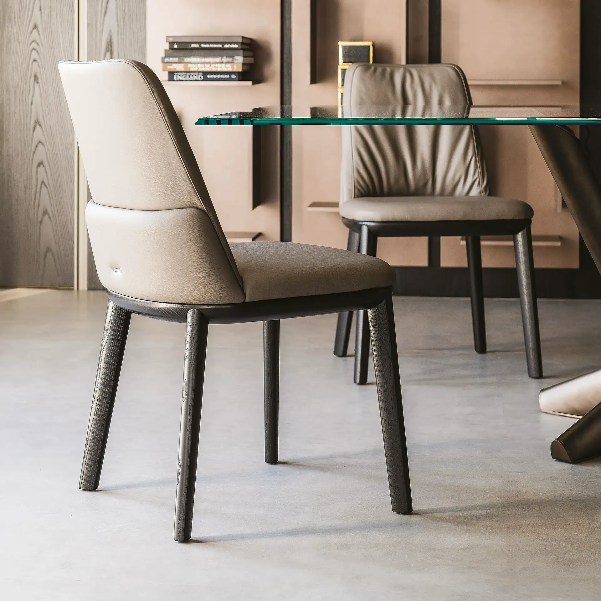 Two modern dining chairs with light beige upholstery and dark wooden legs, positioned around a clear glass table. The background features contemporary wall paneling and shelves with books.