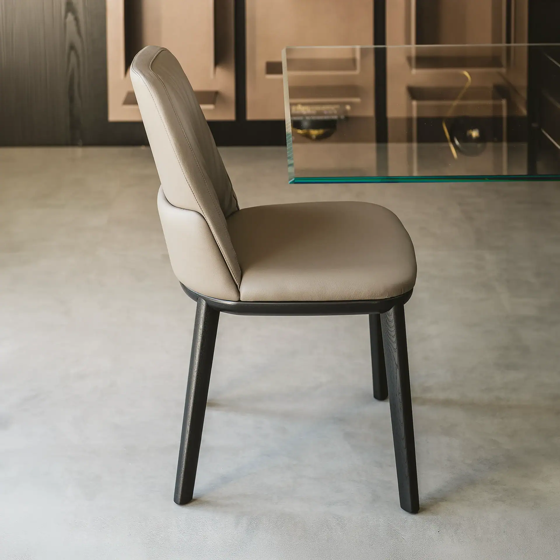 A modern dining chair with light taupe leather upholstery and dark wood legs, set against a light gray floor and a glass table.