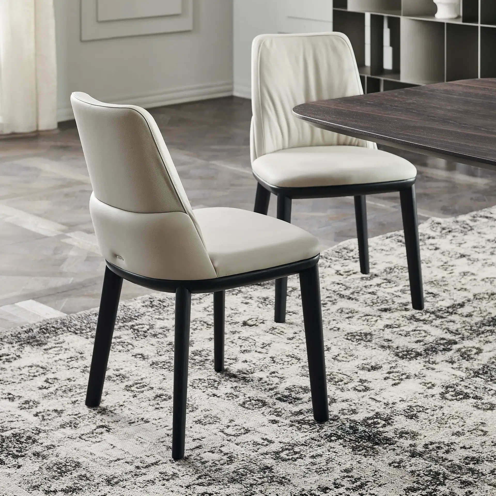 Two light beige upholstered dining chairs with dark legs on a black and white patterned rug, next to a dark dining table.