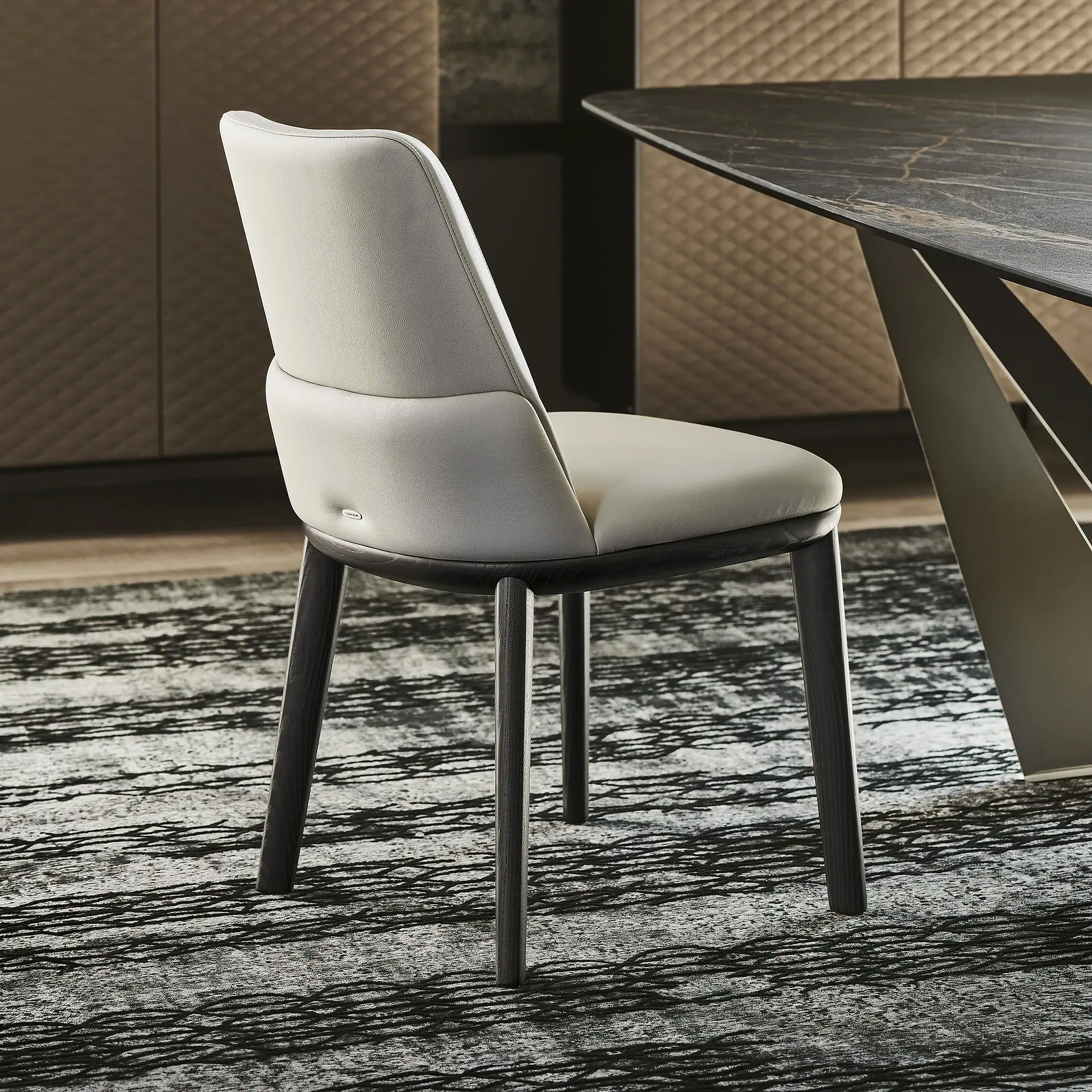 A light beige leather dining chair with dark wooden legs sits on a black and white patterned rug next to a dark marble table.