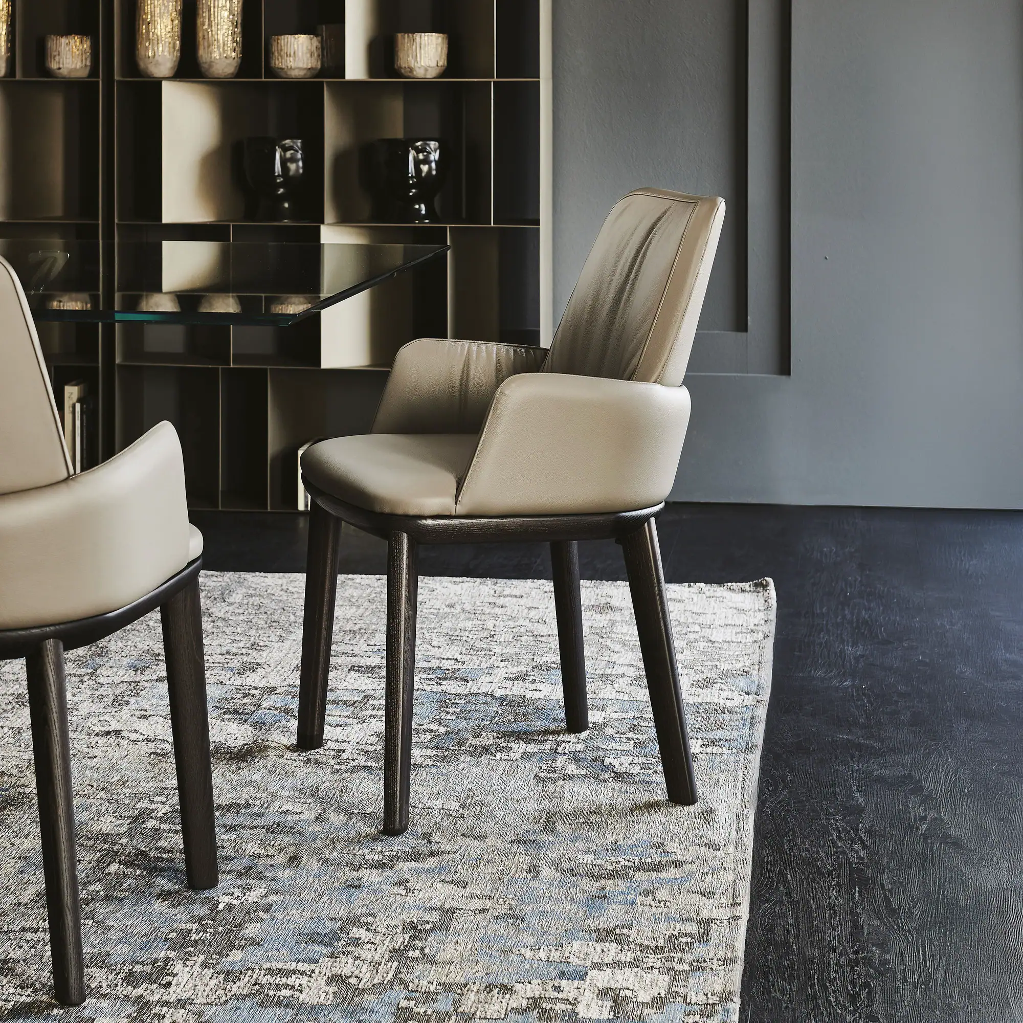 Two modern light beige leather dining chairs with dark wooden legs on an abstract patterned rug, with a dark shelving unit and grey wall in the background.