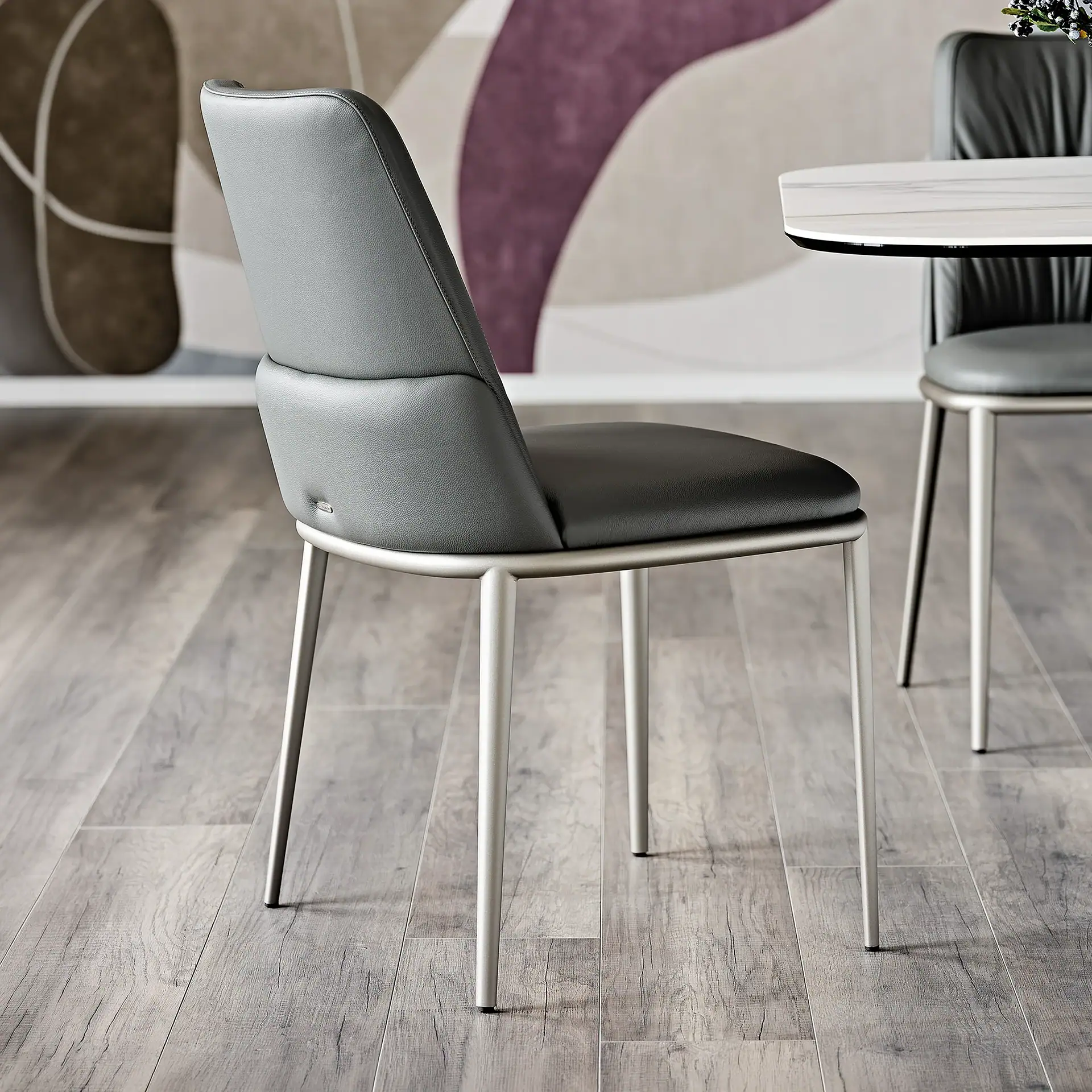 A dark gray upholstered dining chair with brushed metal legs on a light wood-look floor, with an abstract wall and marble-top table in the background.