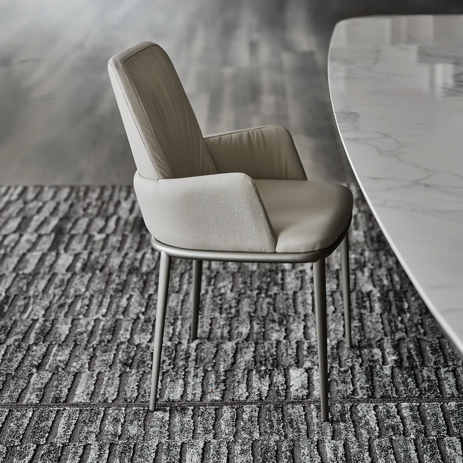 A light-colored modern dining chair with a metal frame sits on a dark gray textured rug next to a white marble table.
