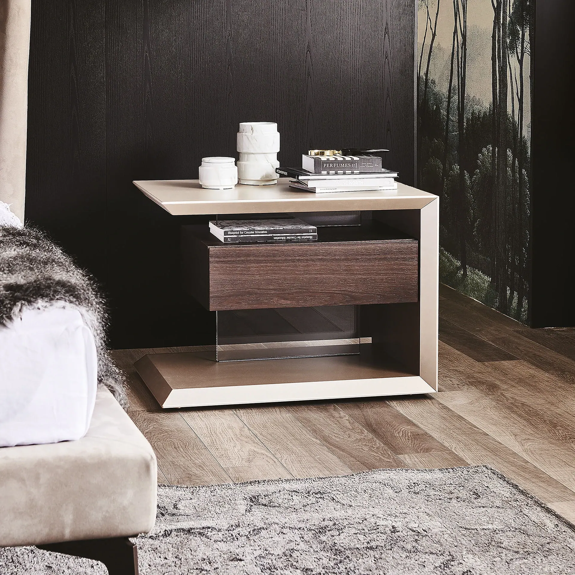 A modern bedside table with a light beige frame and dark wood drawer, adorned with white vases and books, next to a bed with a furry throw. A dark forest mural covers the wall behind it.