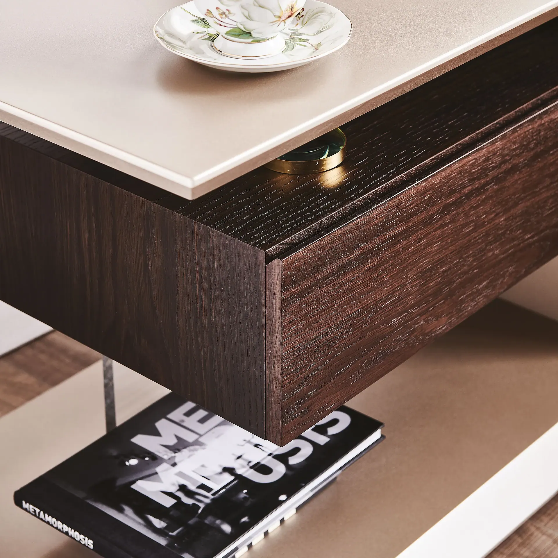 Close-up of a modern side table with a light beige top, a floral teacup, a dark wood drawer, and a black book titled 'METAMORPHOSIS' on a lower shelf.