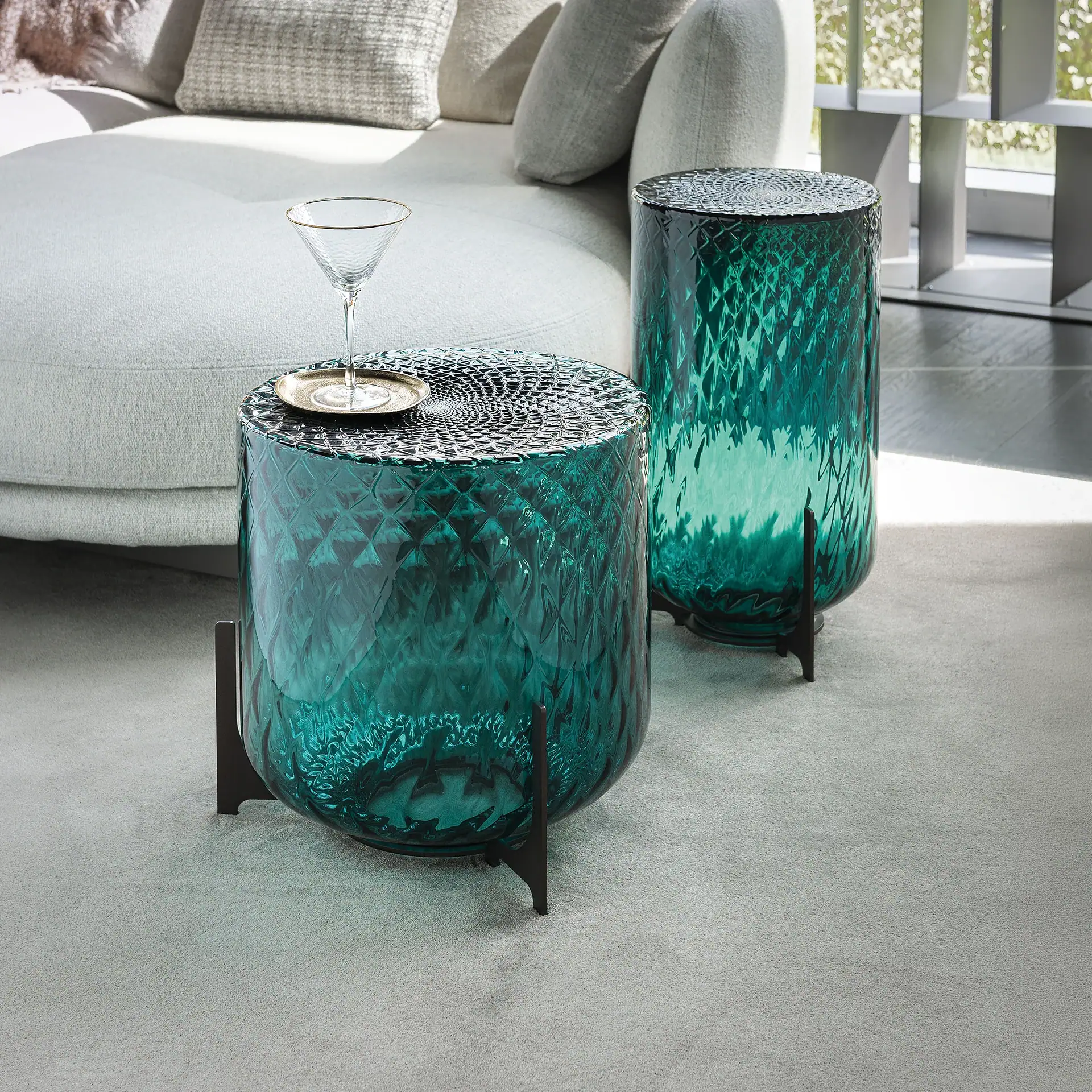 Two cylindrical teal glass side tables on a light grey carpet. A martini glass sits on the smaller table, with a light-colored sofa in the background.