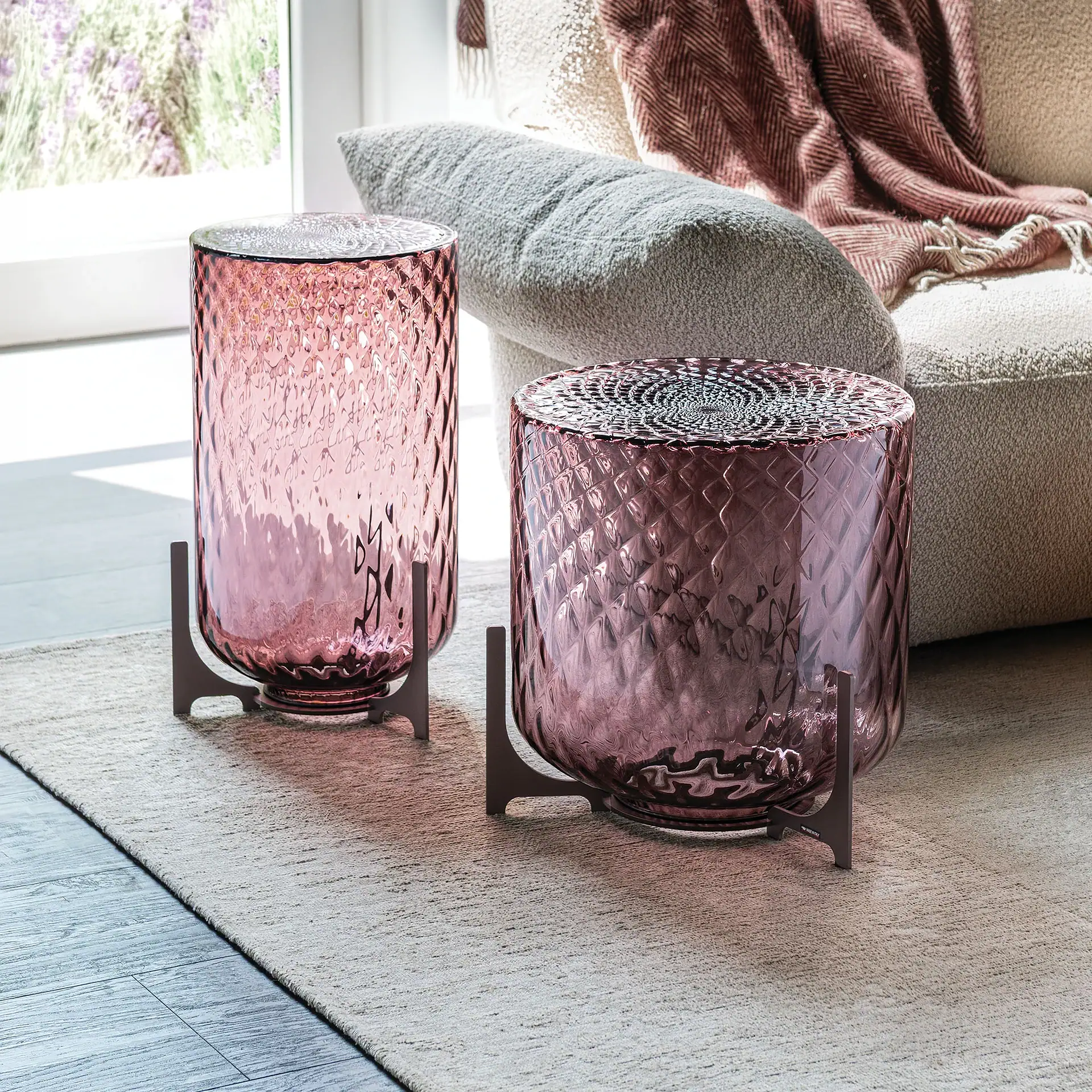 Two textured pink glass cylindrical decor pieces on dark metal stands, placed on a rug in front of a sofa with a throw blanket.