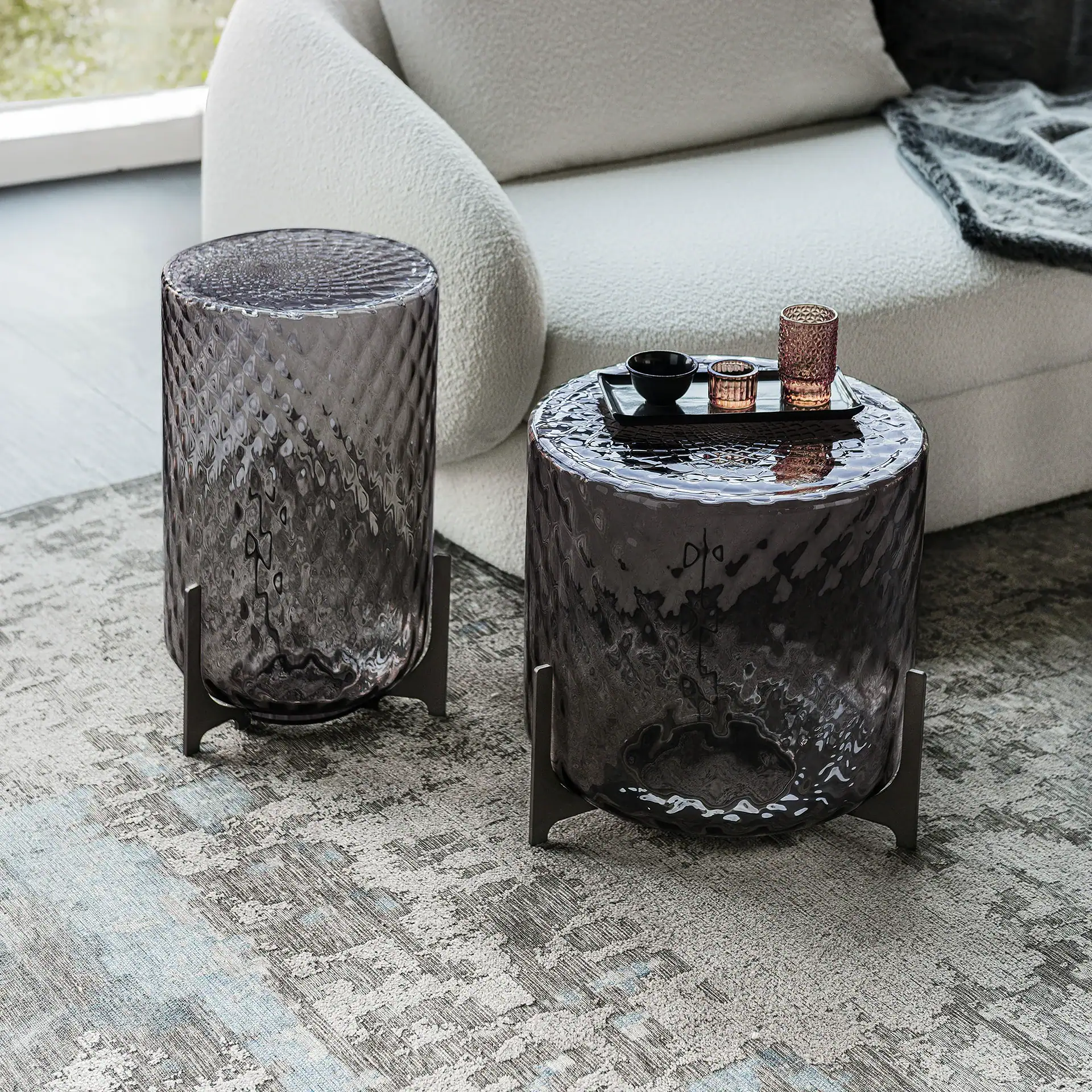 Two dark, textured glass cylindrical side tables with metal bases on a patterned rug next to a cream sofa. One table holds a tray with a black bowl and two votives.