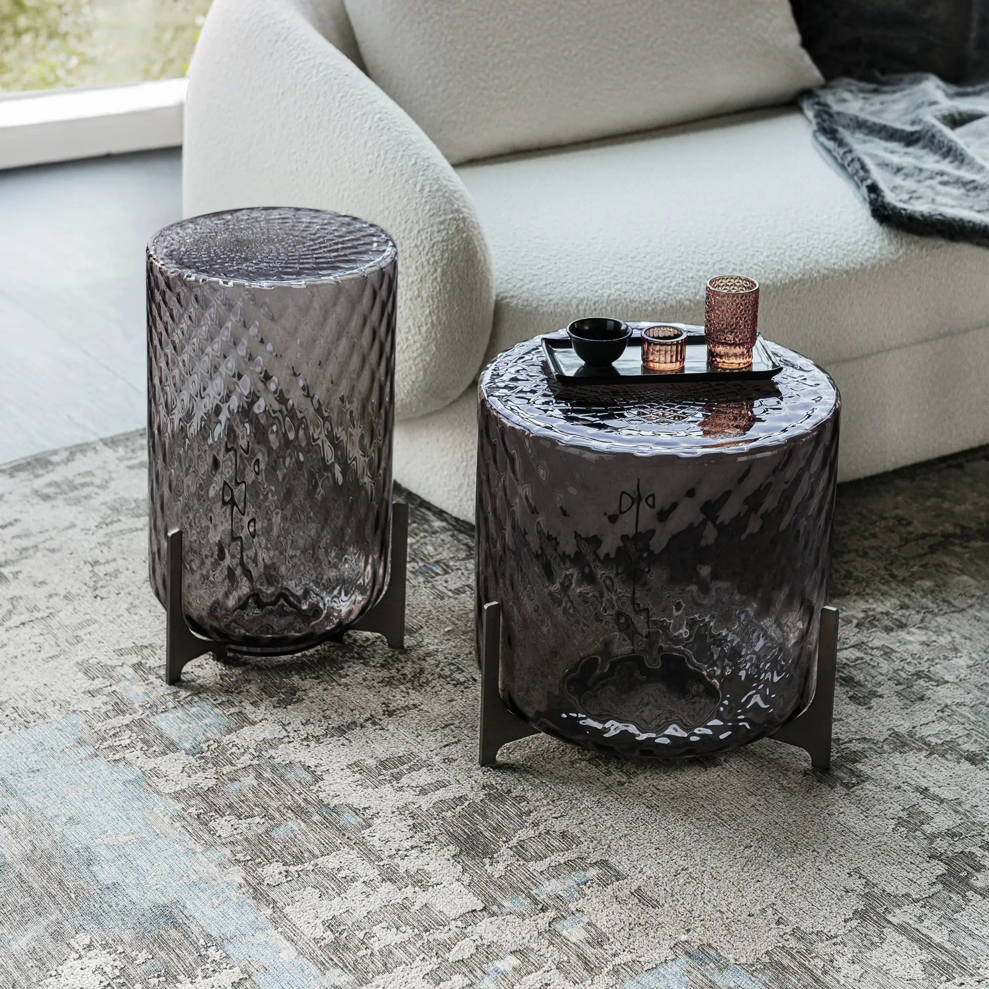 Two dark, textured glass cylindrical side tables with metal bases on a patterned rug next to a cream sofa. One table holds a tray with a black bowl and two votives.