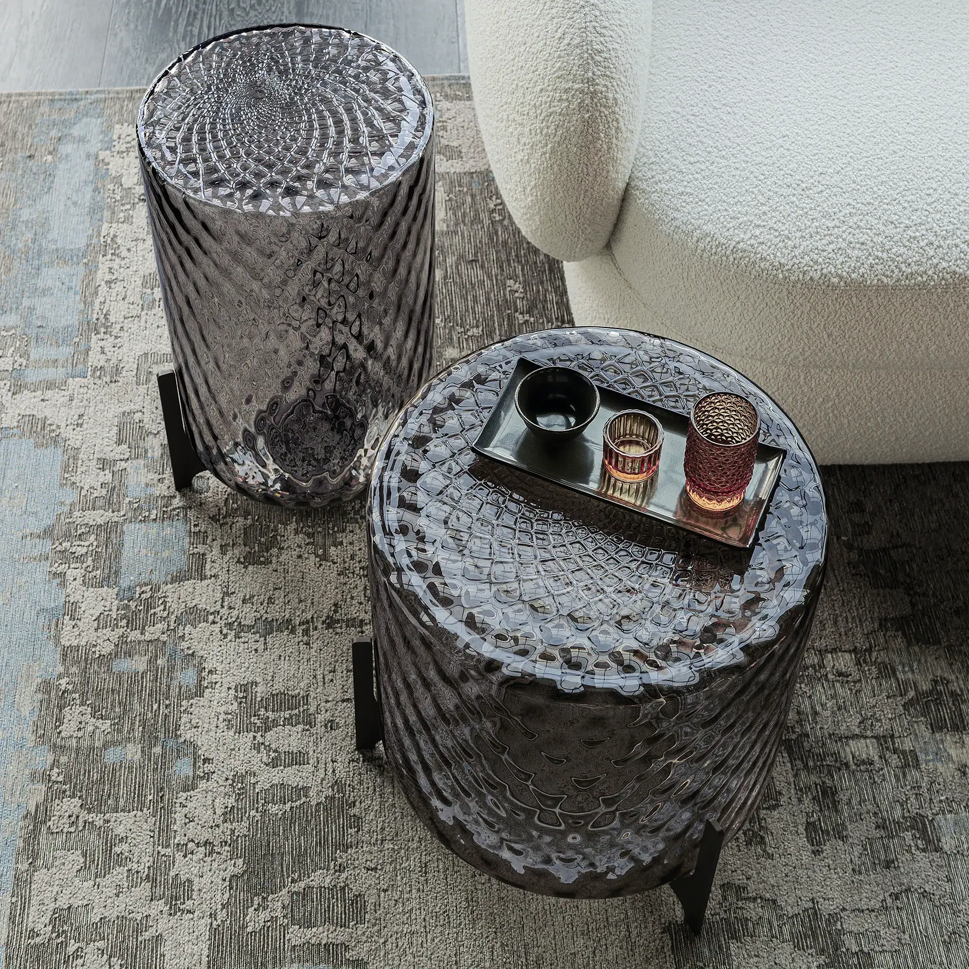Two dark, textured glass side tables next to a white bouclé sofa on an abstract grey and blue rug. One table holds a tray with a bowl and two votive candle holders.