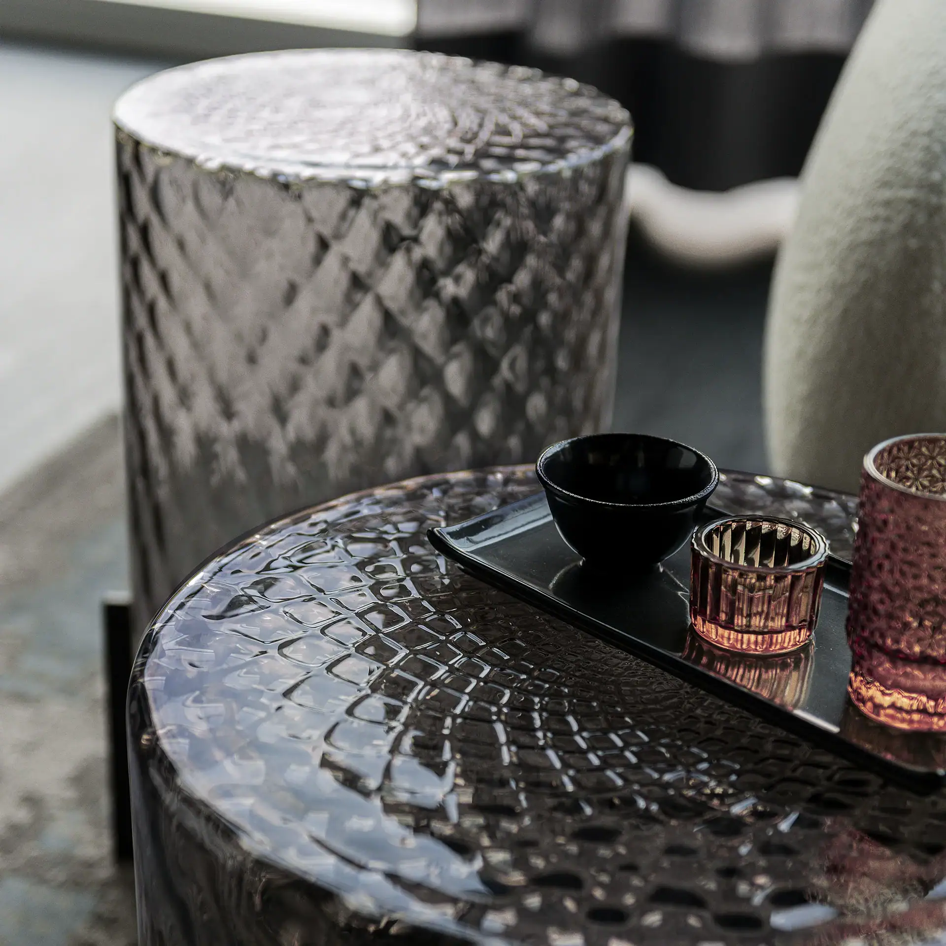 Two textured, reflective cylindrical side tables. The front table holds a black tray with a black bowl and two amber glass candle holders.