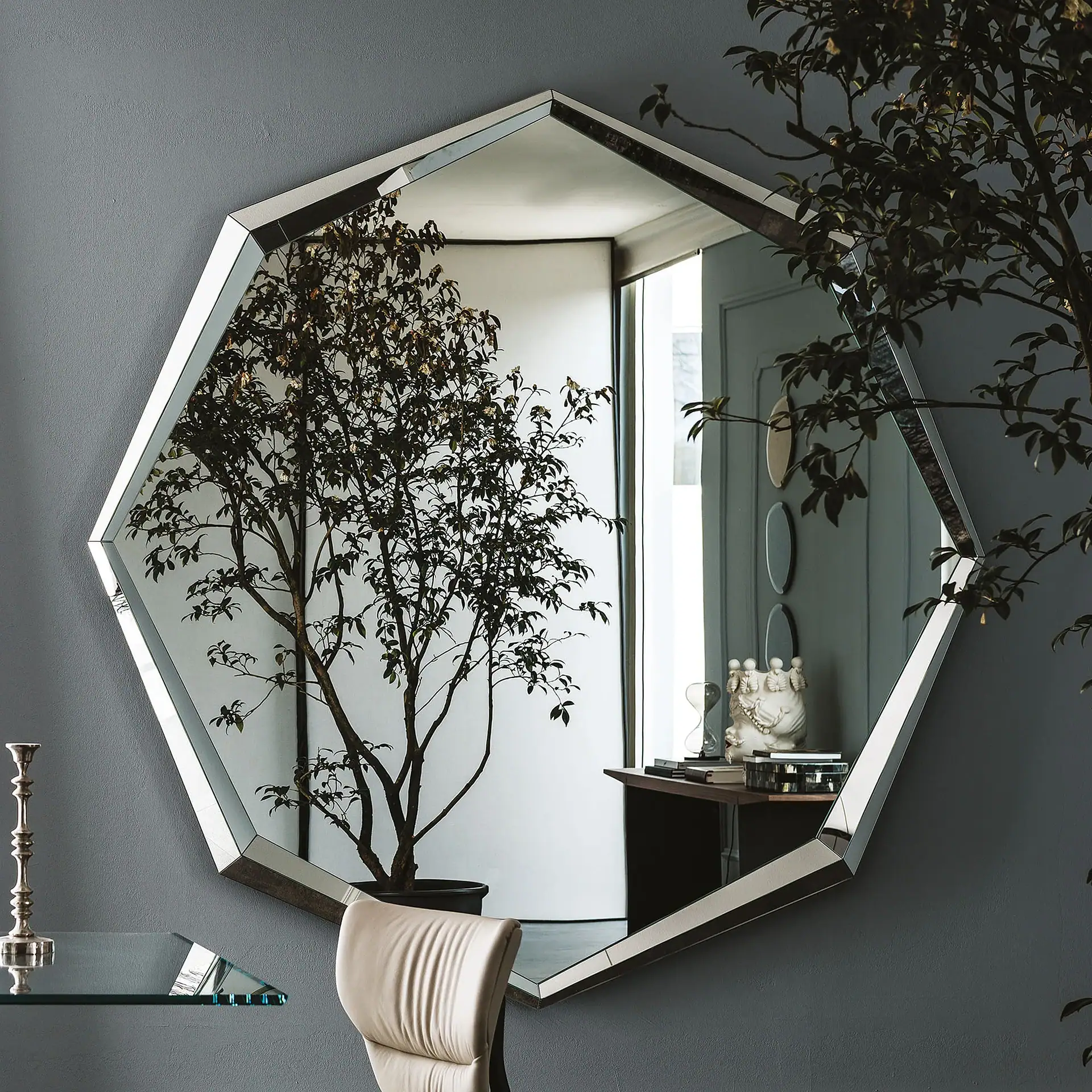 An octagonal mirror with a faceted frame on a gray wall reflects a room with a potted plant, a console table with decor, and a window. A silver candlestick is on a glass shelf to the left.
