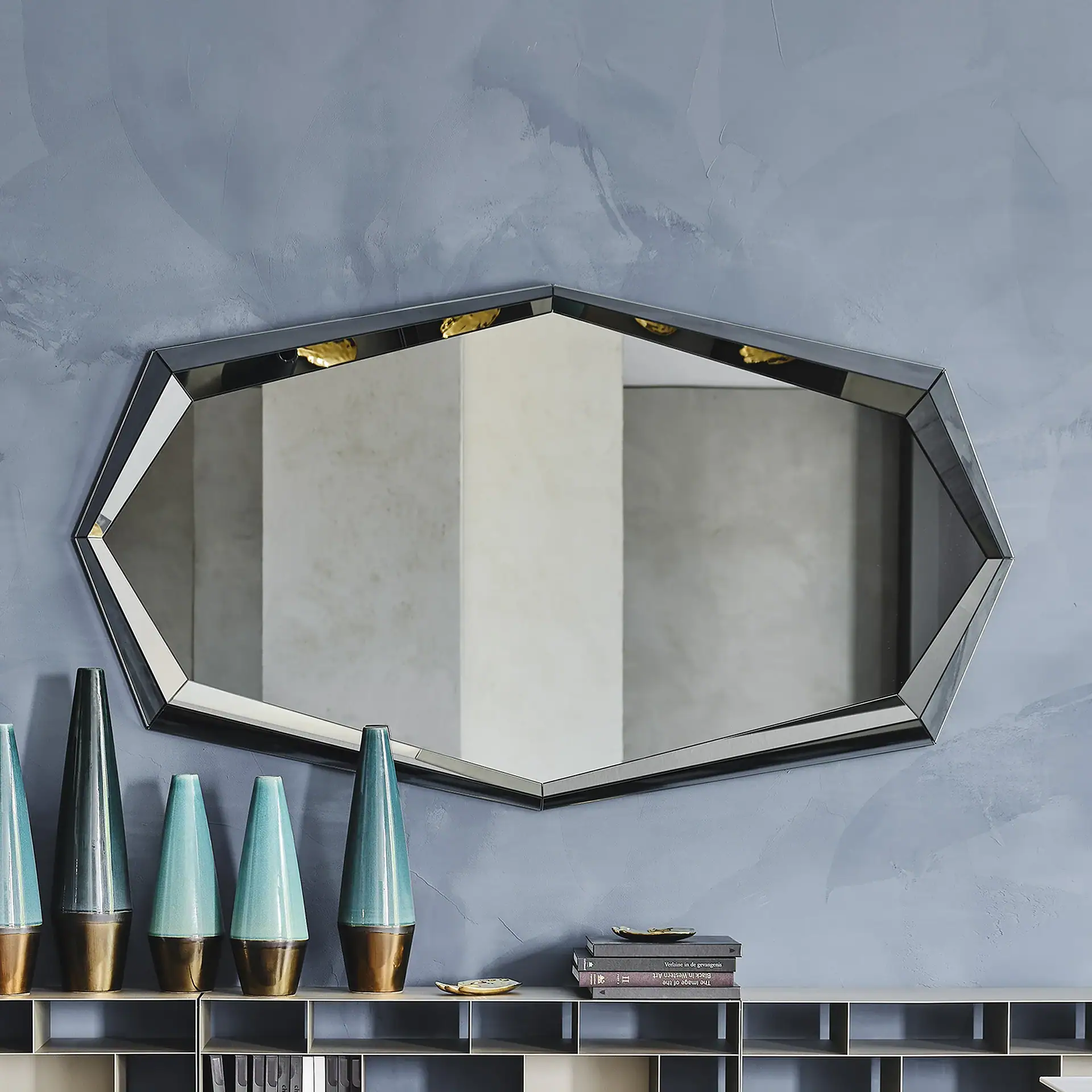 A large, dark-framed, irregular hexagonal mirror hangs on a textured blue-grey wall above a console table with five teal and bronze conical vases and a stack of books.