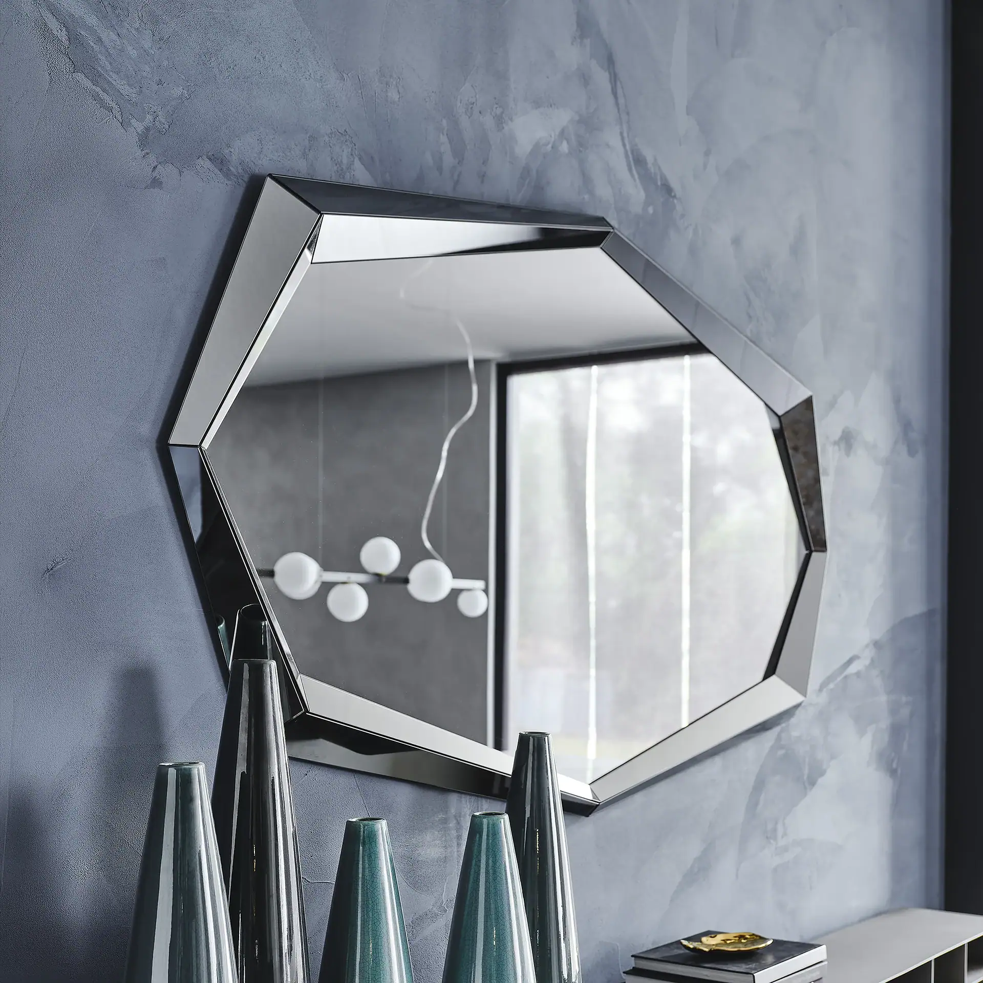 A geometric mirror with a beveled frame on a textured gray wall, reflecting a modern chandelier and a window. Below it are cone-shaped vases and books with a gold dish.