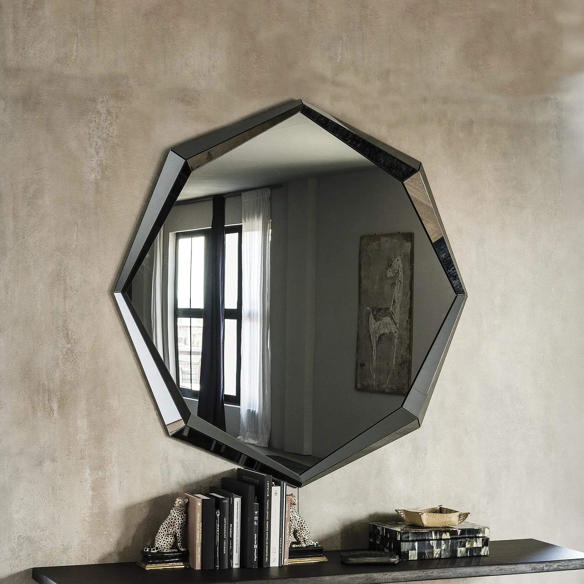 An octagonal mirror with a dark, faceted frame hanging on a textured beige wall above a dark console table. The mirror reflects a window and artwork. The table holds books, leopard figurines, and decorative boxes.