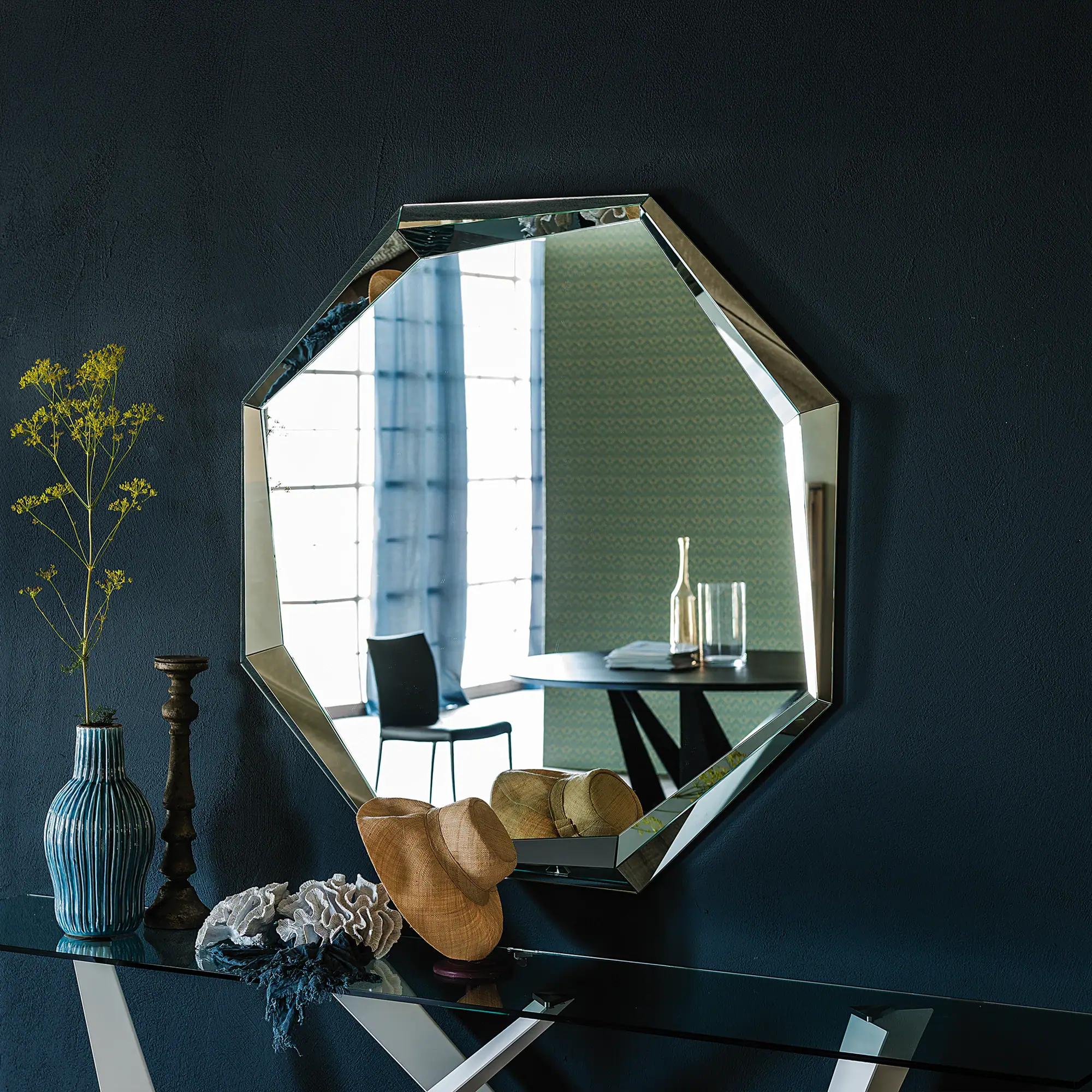 A modern octagonal mirror hangs above a glass console table with a blue vase, yellow plant, candlestick, coral decor, and a woven hat, all against a dark blue wall.